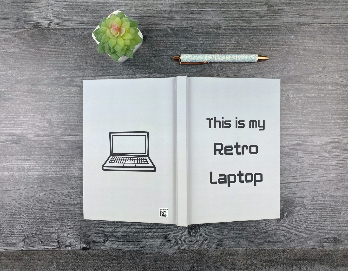 Hardcover journal open to show both covers, one with a simple hand-drawn laptop illustration, the other reading "This is my retro laptop" in a pixelated font, resting on a grey wood surface next to a glitter pen and small succulent.
