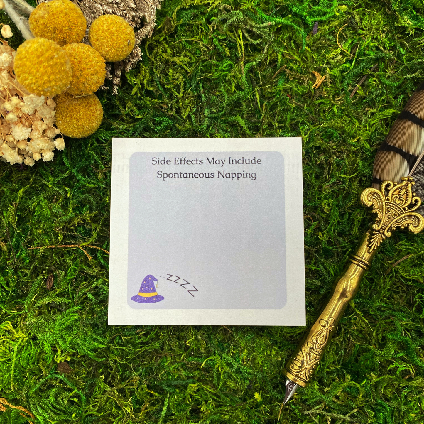 3x3 inch sticky note pad reading "Side Effects May Include Spontaneous Napping" with purple witch hat and zzz illustrations on soft gray background, displayed on green moss with yellow dried flowers and vintage gold quill pen.