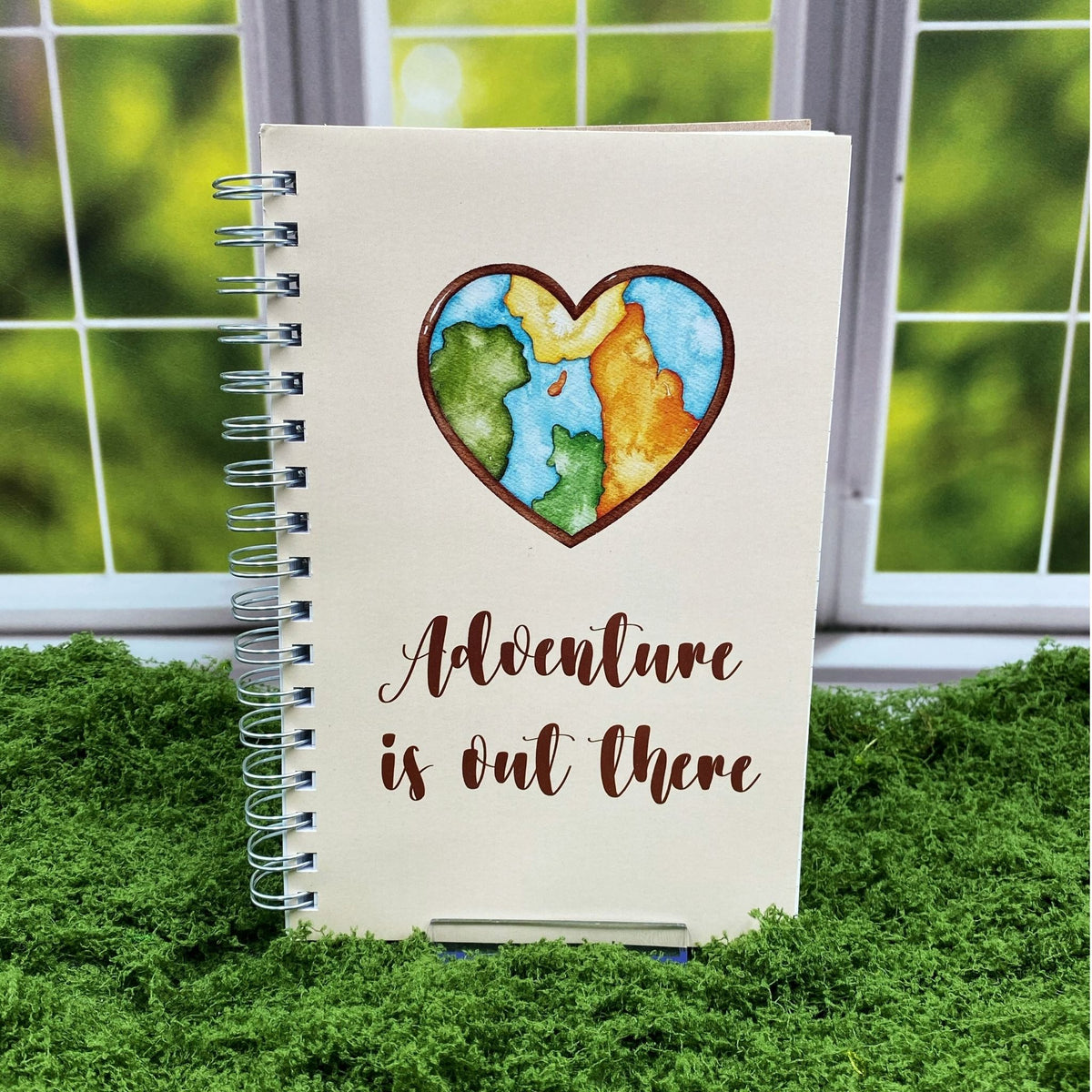 Spiral-bound cream notebook with a watercolor heart-shaped world map illustration above text reading 'Adventure is out there,' displayed on a moss mat with a window backdrop.