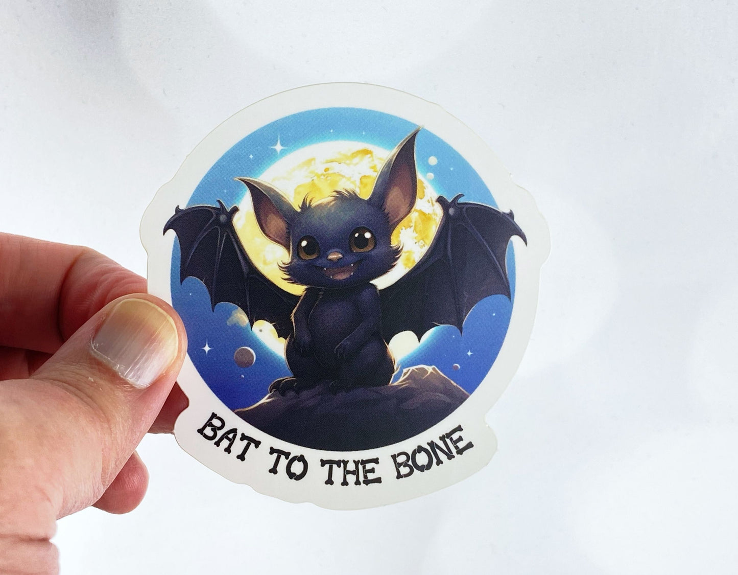 A hand holds a sticker illustration of a wide-eyed baby bat perched on a rock under a full moon, reading "Bat to the bone," against a white background.