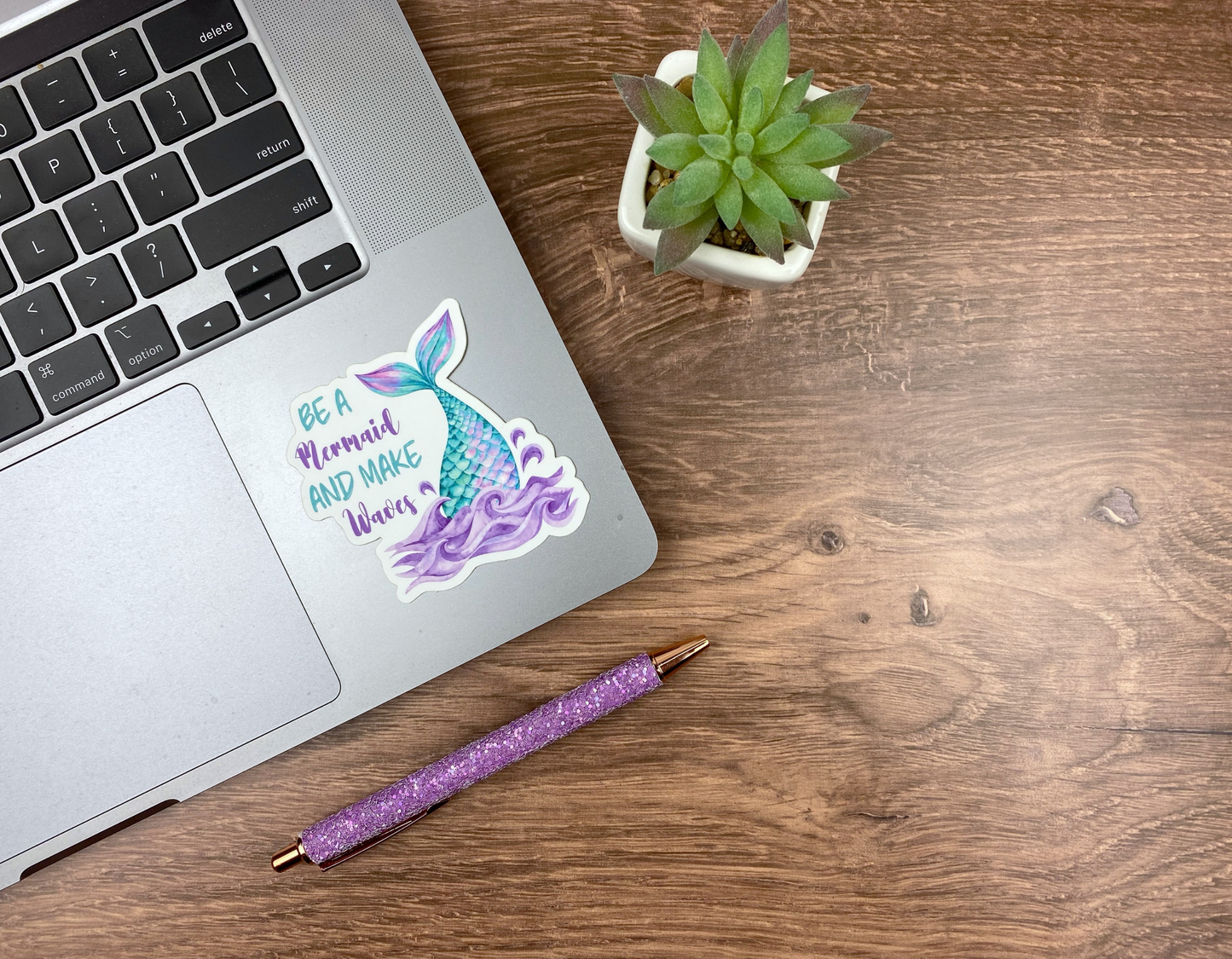 Mermaid tail and waves sticker applied to the corner of a silver laptop trackpad, next to a succulent and a glitter pen on a wood desk