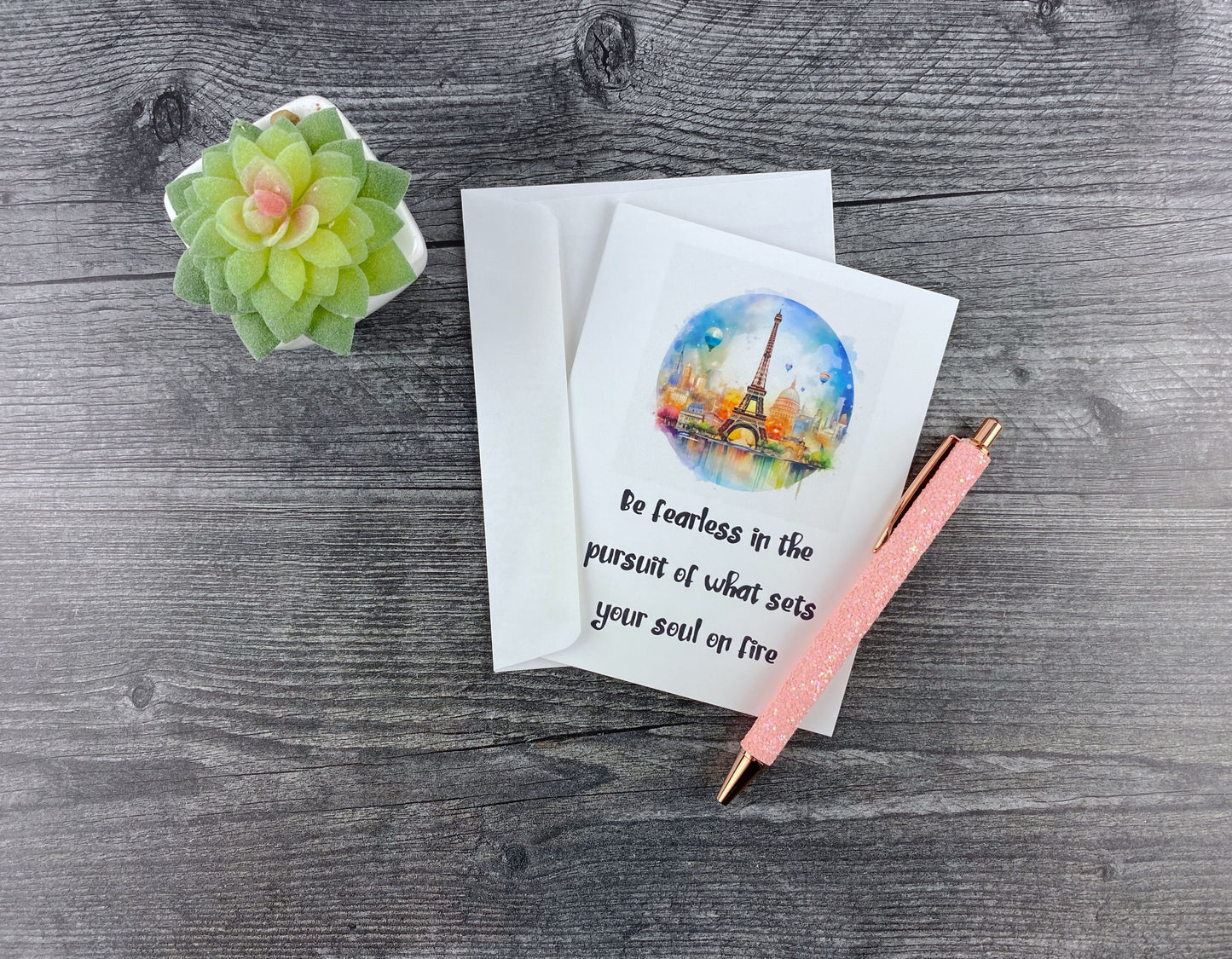 Greeting card and matching envelope, front featuring a circular watercolor illustration of the Eiffel Tower and hot air balloons, reading "Be fearless in the pursuit of what sets your soul on fire," resting on a grey wood surface next to a pink glitter pen and small succulent.