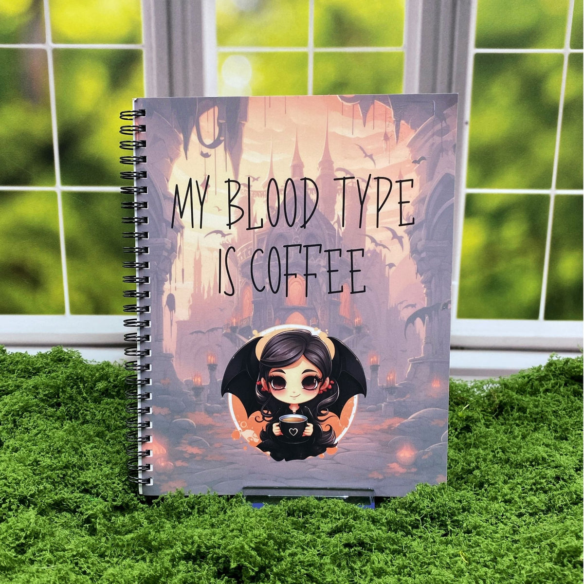 Spiral-bound notebook with a gothic castle illustration and a chibi vampire holding a coffee cup, reading 'My blood type is coffee,' displayed on a moss mat with a window backdrop.