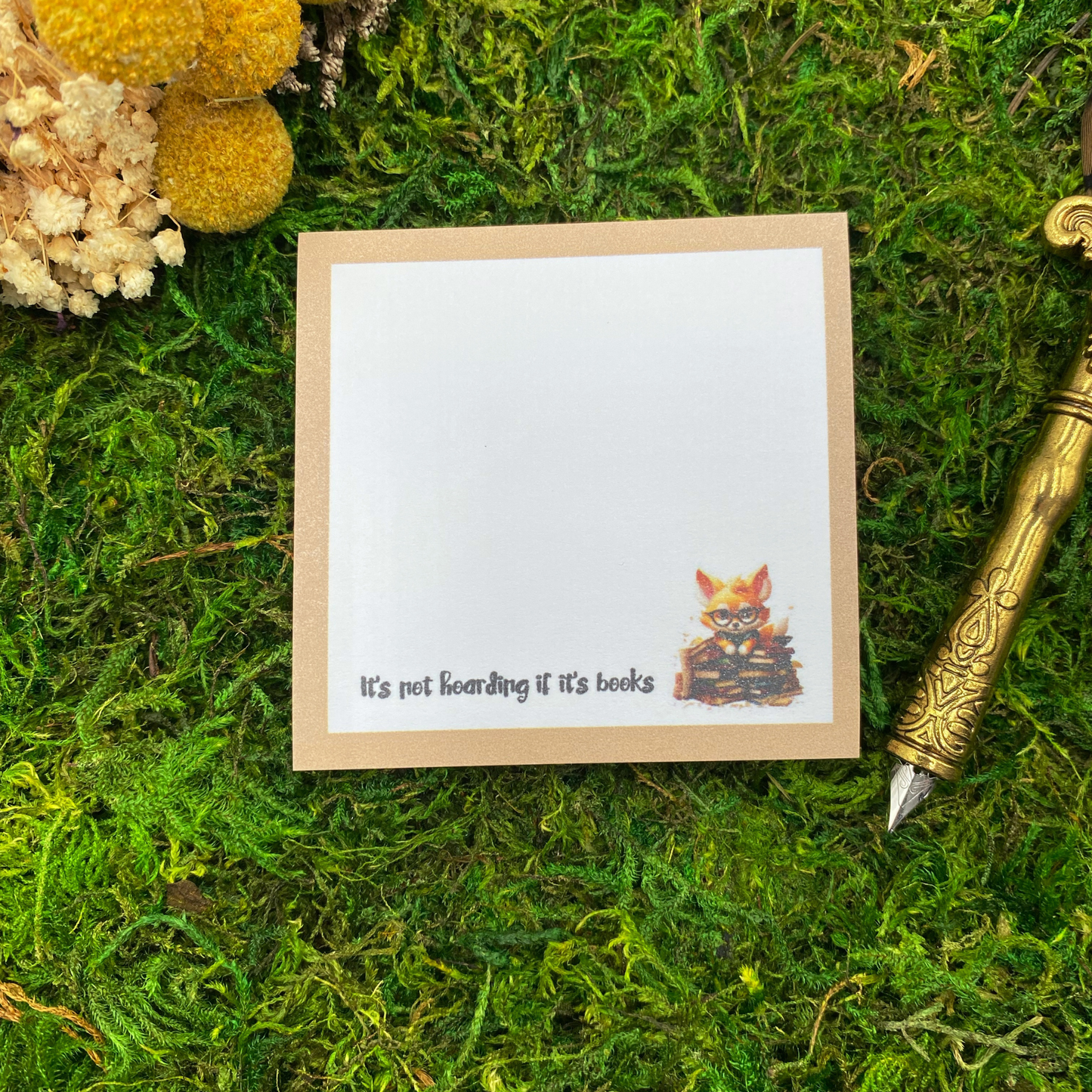 Square sticky note pad with a tan border, a fox wearing glasses sitting on a stack of books, and the words "It's not hoarding if it's books," resting on moss next to dried yellow and white flowers and a gold quill pen.