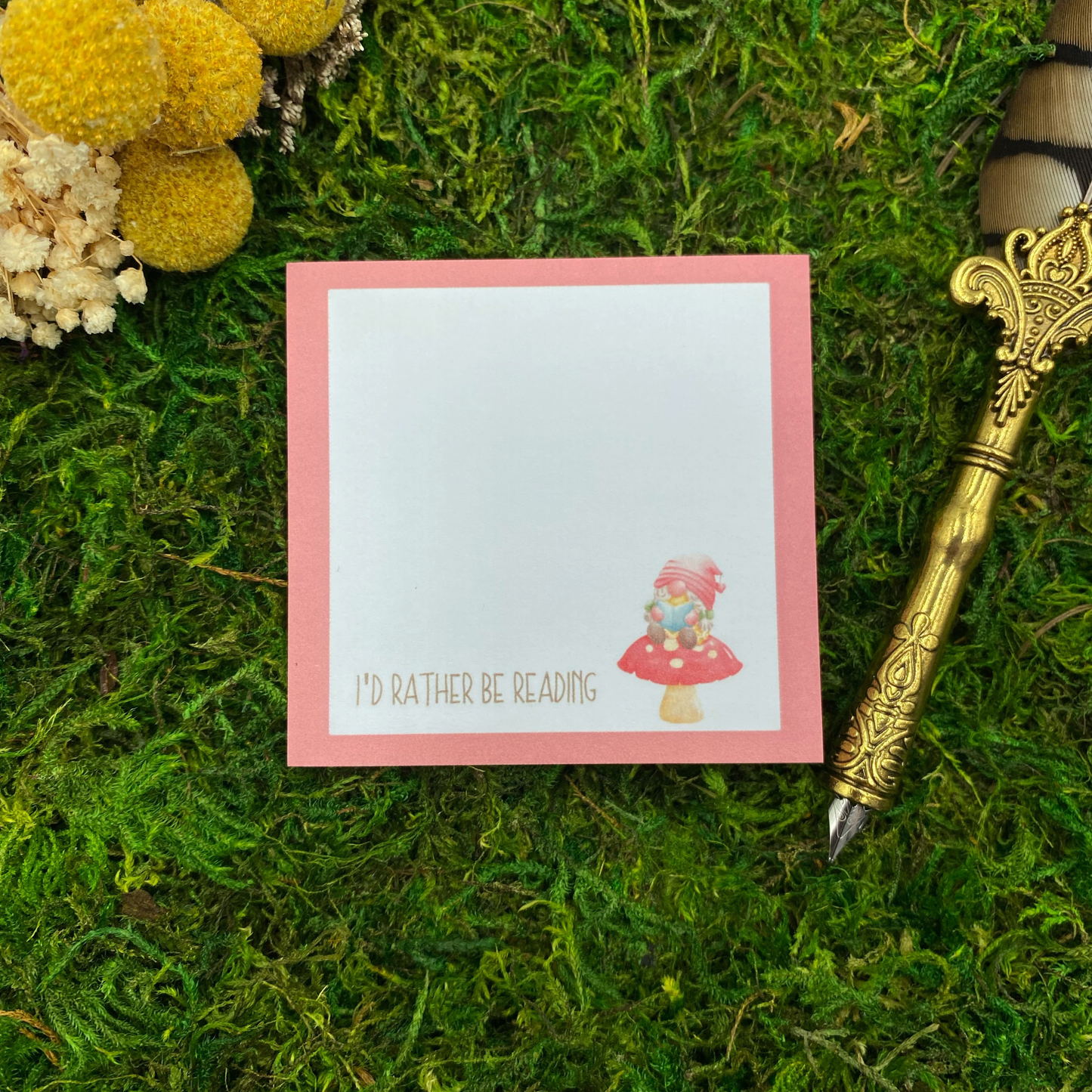 Square sticky note pad with a pink border, a gnome sitting on a red mushroom, and the words "I'd rather be reading," resting on moss next to dried yellow and white flowers and a gold quill pen.