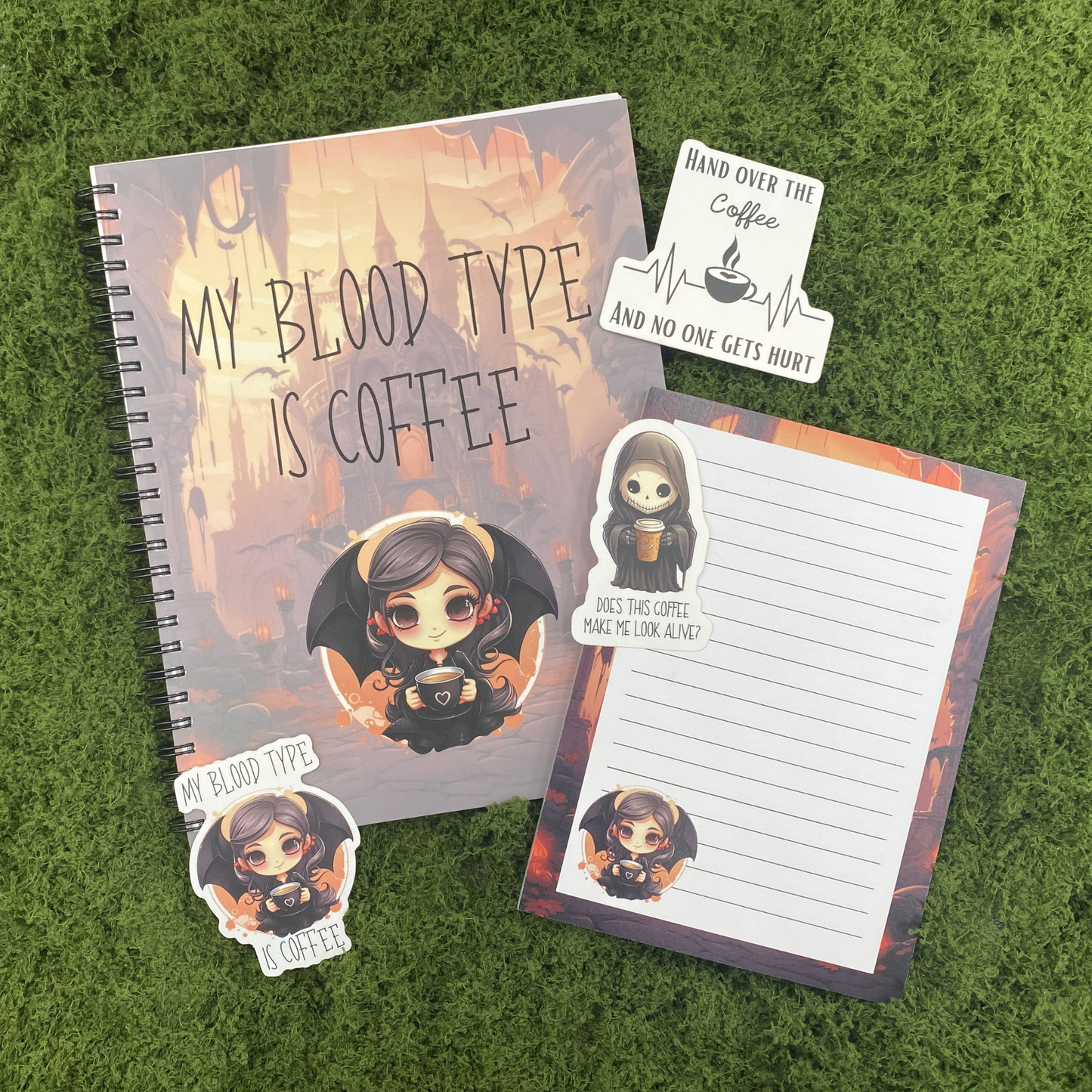 A gothic desk gift set including a my blood type is coffee spiral notebook, a matching 5x7 lined notepad, and three die-cut stickers featuring a vampire girl, a grim reaper with coffee, and a hand over the coffee heartbeat monitor, arranged on a green moss surface