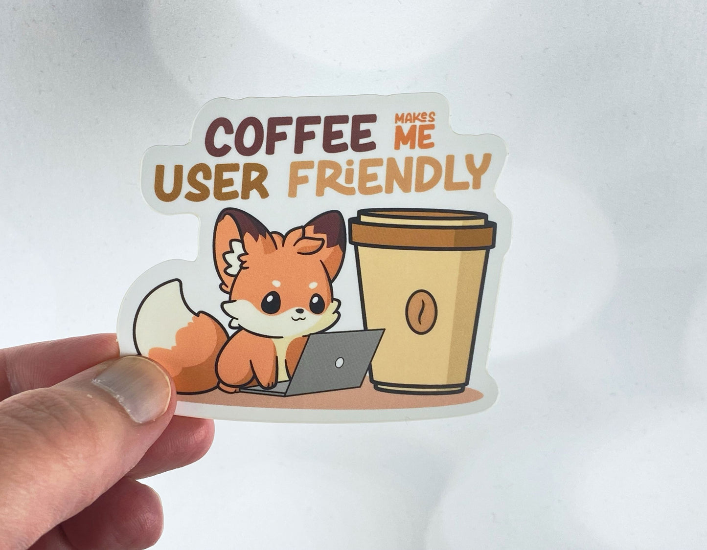 Die-cut vinyl sticker held between fingers, showing a fox at a laptop next to a large coffee cup, with the text "Coffee makes me user friendly," on a white background
