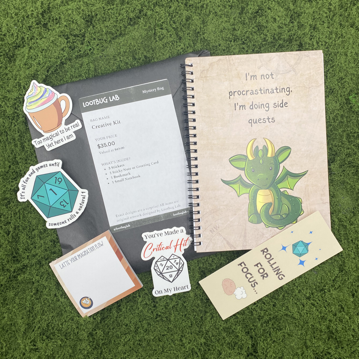 A Lootbug Lab creative kit mystery bag with a reveal card surrounded by sample items including stickers, a bookmark, a small notebook, and a sticky note pad on a green moss surface