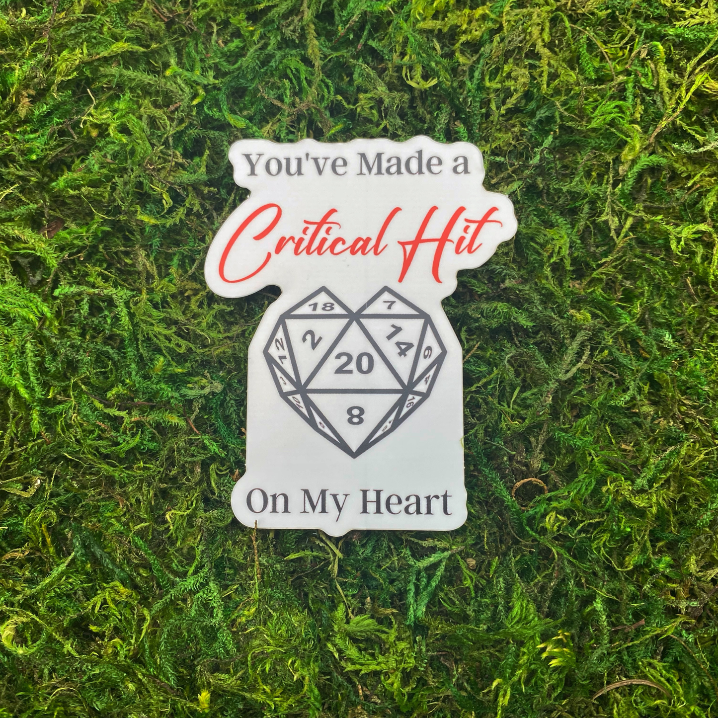 A sticker reading "You've made a critical hit on my heart" with a heart-shaped twenty-sided die showing the number 20, resting on artificial moss.