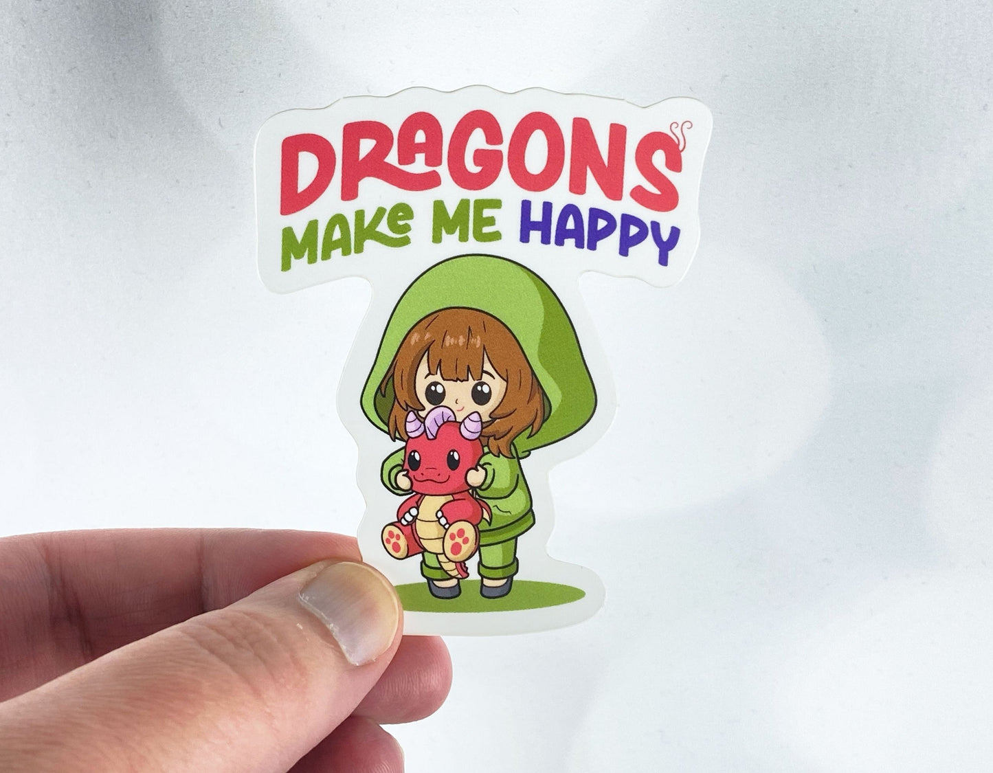 Die-cut vinyl sticker held between fingers, showing a girl in a green hoodie holding a red baby dragon, above the text "Dragons make me happy," on a white background
