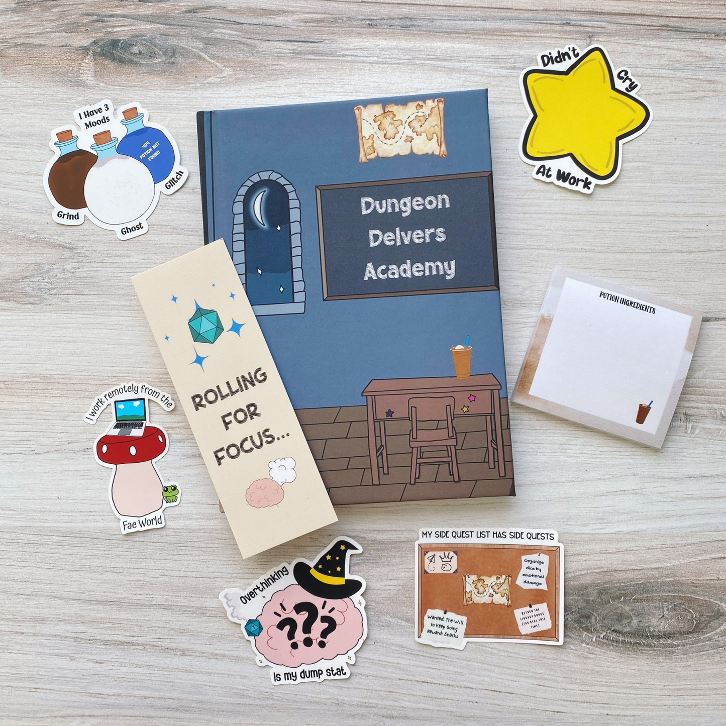 A curated gift set laid out on a light wood background, including a spiral notebook reading "Dungeon Delvers Academy," a bookmark reading "Rolling for Focus," a sticky note pad reading "Potion Ingredients," and five tabletop RPG and work-humor stickers