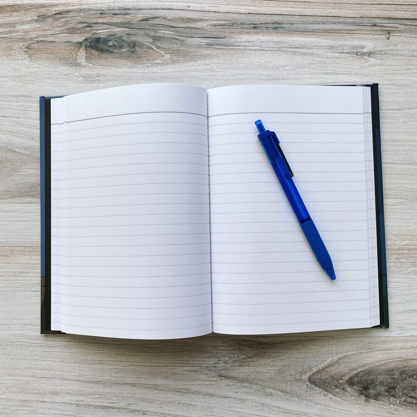Open journal showing blank lined pages with a blue pen resting across them, on a wood surface.