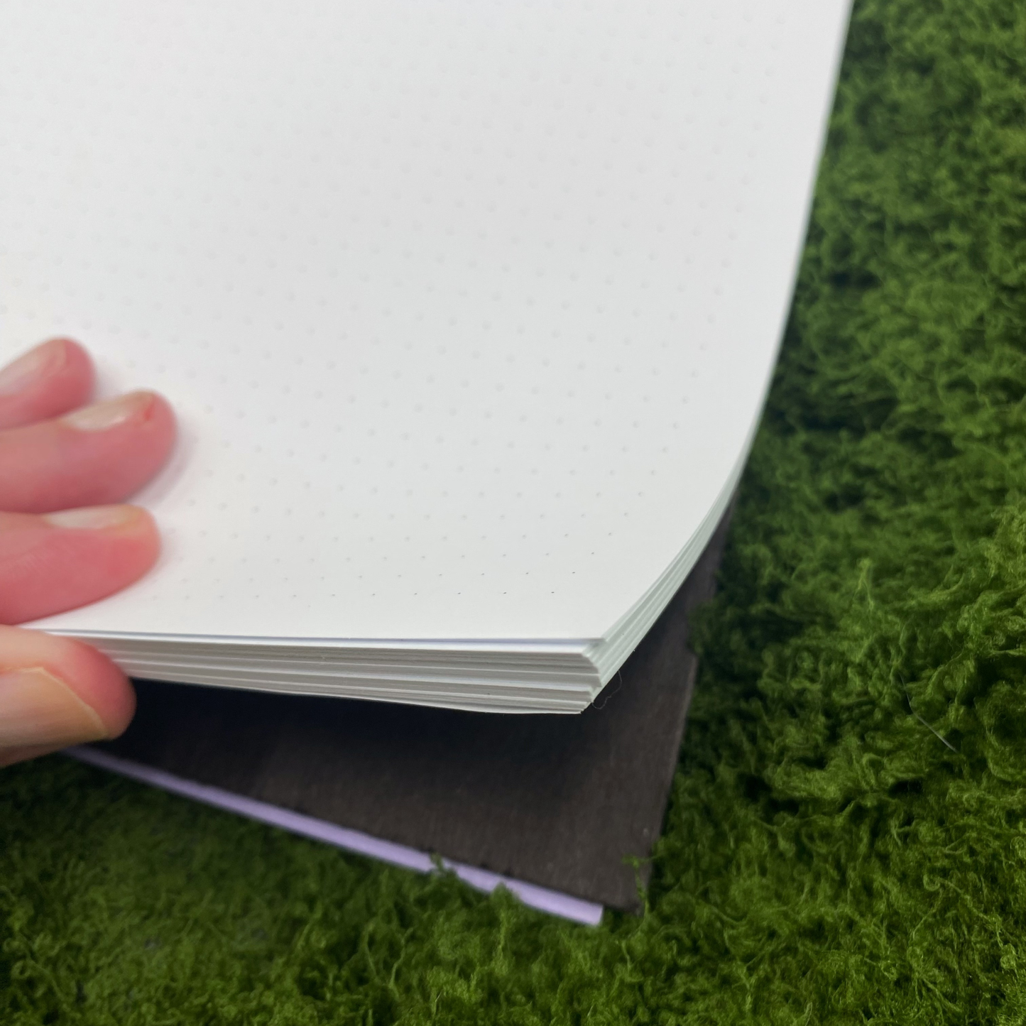 Close-up of dot grid pages inside a notebook with a lavender-edged cover peeking out, shown on an artificial grass surface.