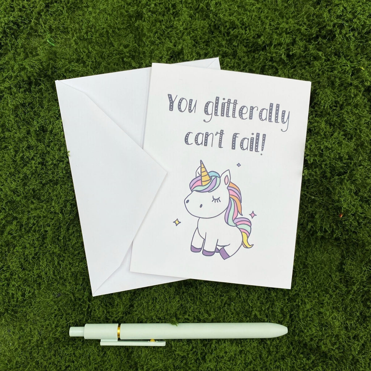 "You Glitterally Can't Fail!" greeting card lying on a white envelope on grass, with a mint green pen beside it for scale.