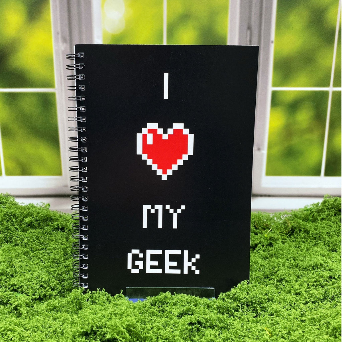 Spiral-bound black notebook reading 'I ♥ My Geek' in pixel-style text with a red pixel heart graphic, displayed on a moss mat with a window backdrop.