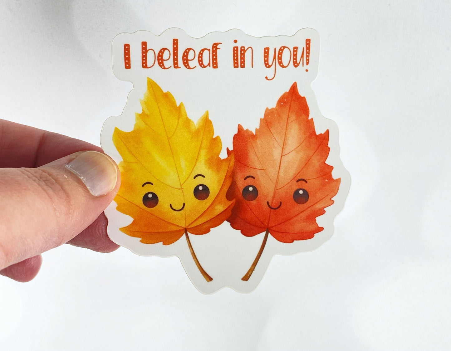A hand holds a sticker illustration of two smiling watercolor autumn leaves, one yellow and one orange, reading "I beleaf in you!" against a white background.