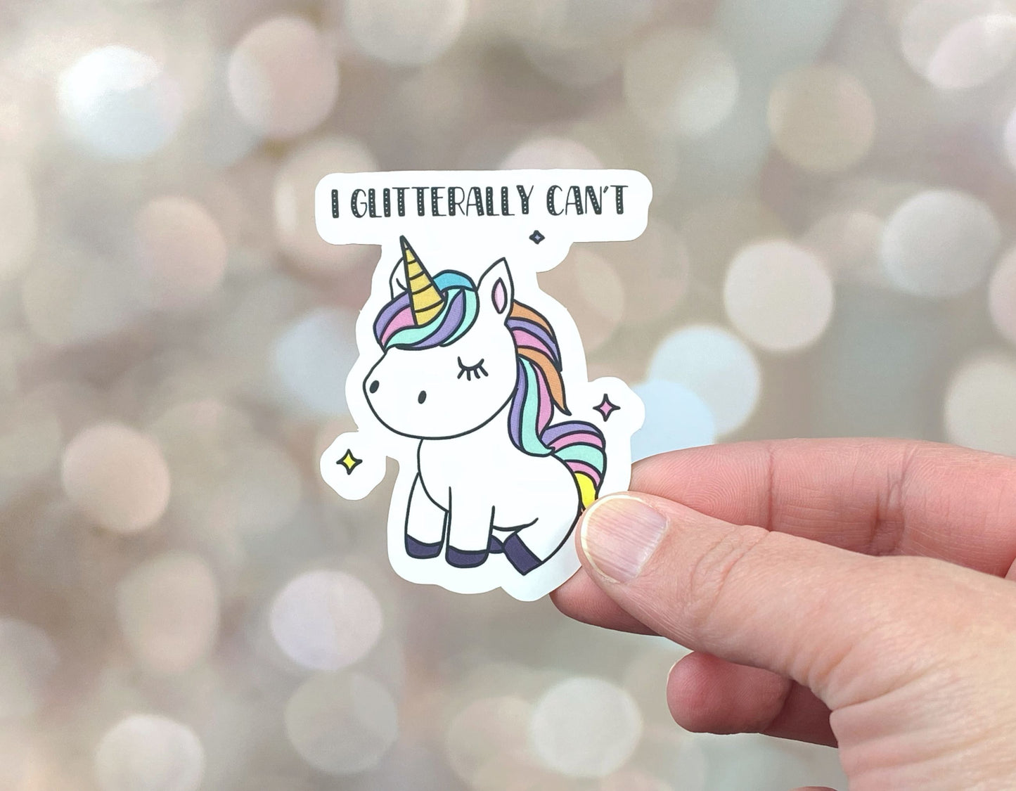 Die-cut vinyl sticker showing a white unicorn with a rainbow mane and tail, with the text "I glitterally can't," on a golden bokeh background