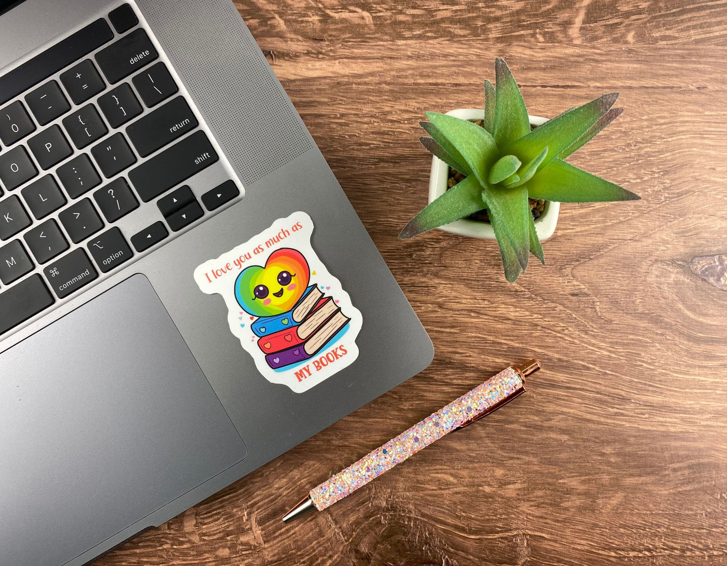 Rainbow heart and book stack sticker applied to the corner of a silver laptop trackpad, next to a succulent and a glitter pen on a wood desk