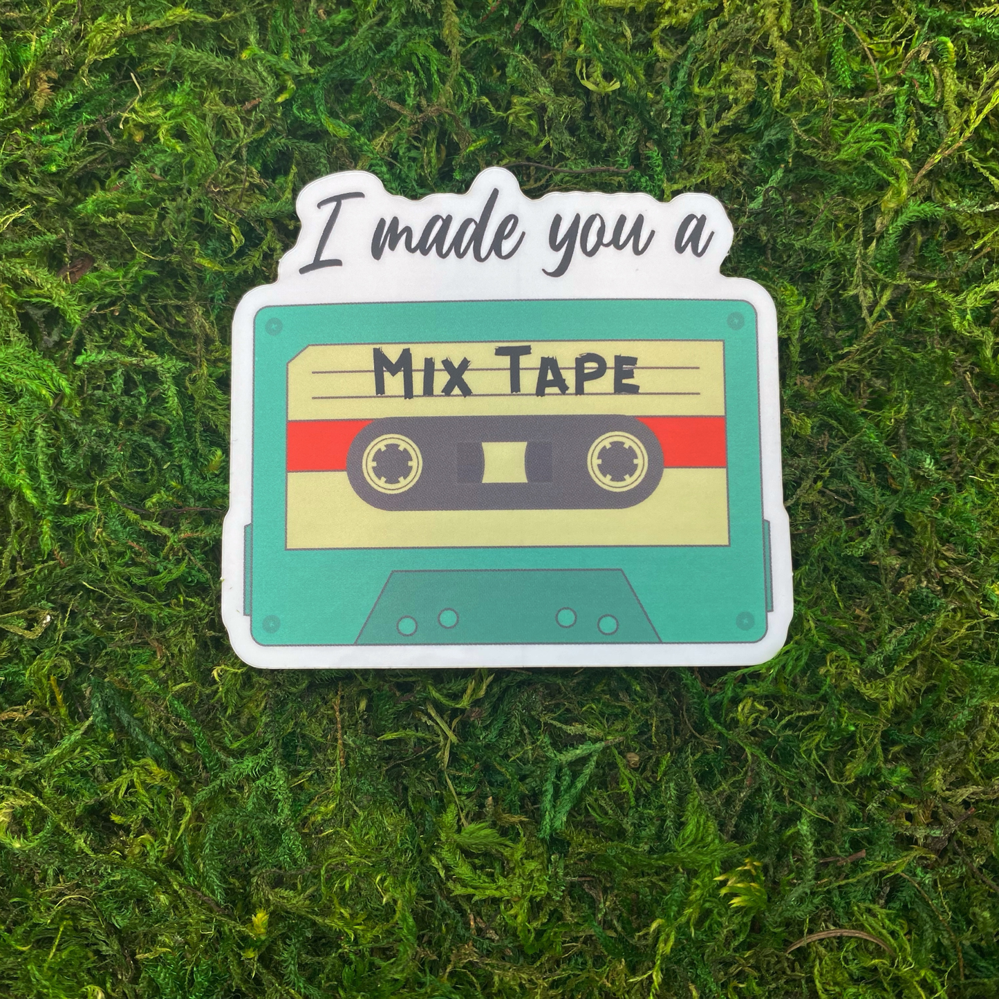A sticker illustration of a teal cassette tape labeled "Mix Tape" with the text "I made you a" above it, resting on artificial moss.