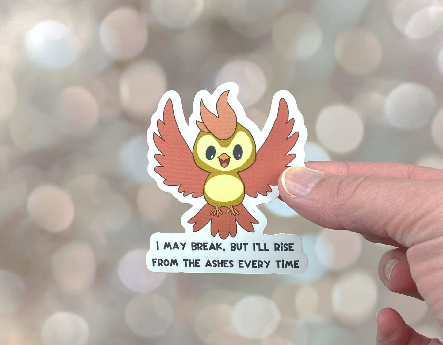 Die-cut vinyl sticker showing an orange and yellow phoenix with wings spread, with the text "I may break, but I'll rise from the ashes every time," on a golden bokeh background