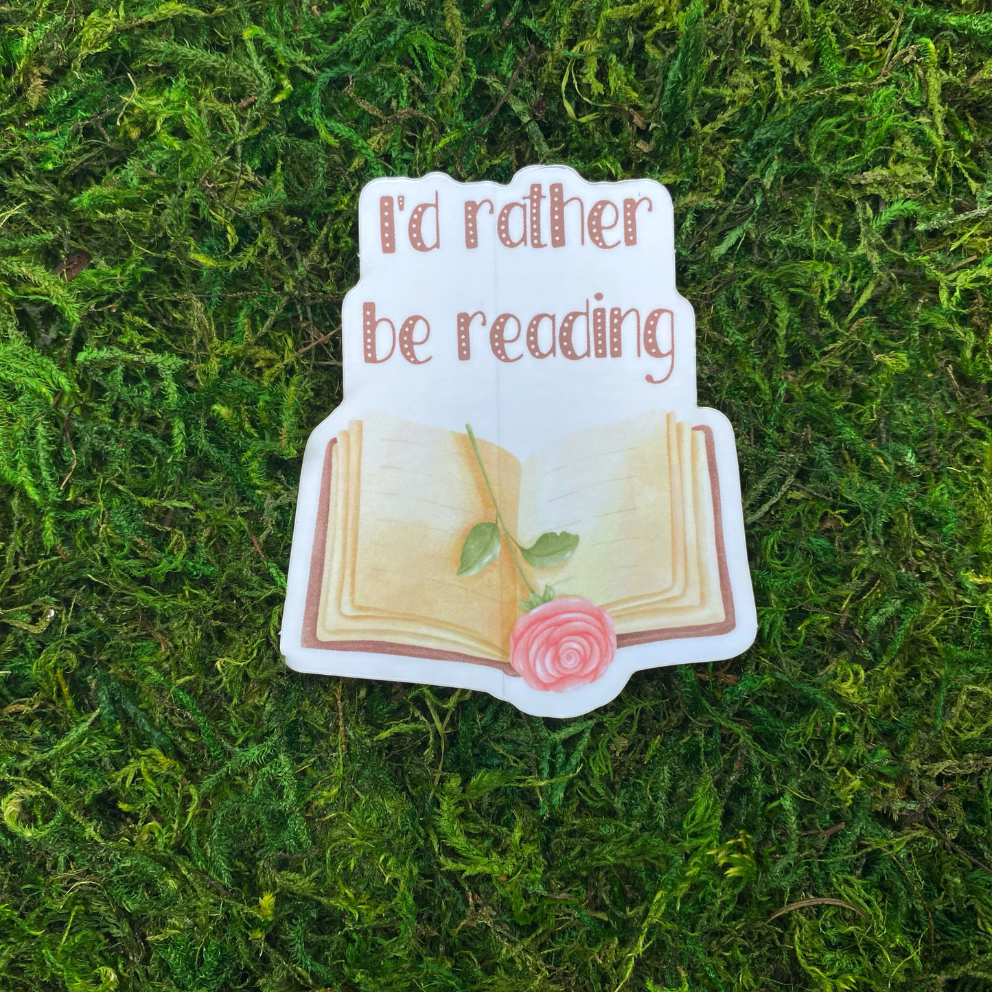 A sticker illustration of an open book with a pink rose resting between the pages, reading "I'd rather be reading," resting on artificial moss.