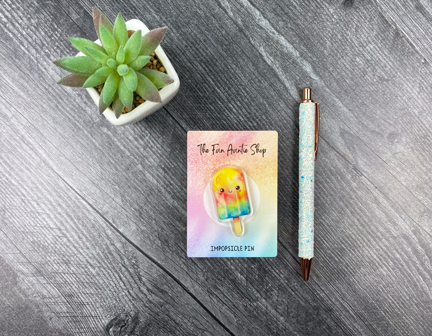 Acrylic pin shaped like a smiling rainbow popsicle on a backing card, next to a succulent and a glitter pen on a gray wood background