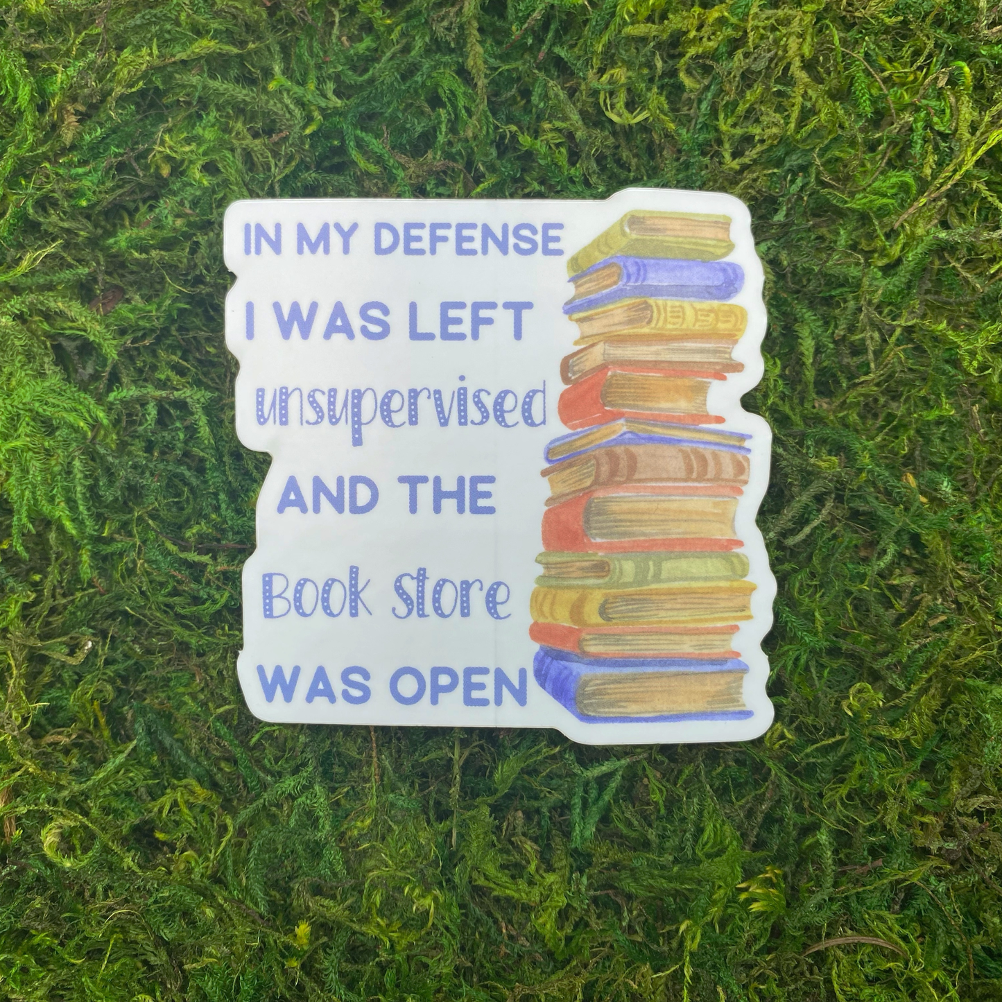 A sticker reading "In my defense I was left unsupervised and the book store was open" beside an illustration of a tall stack of books, resting on artificial moss.