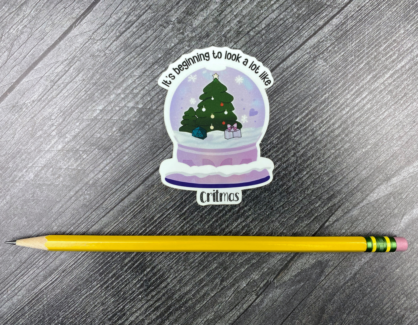 Die-cut vinyl sticker showing a snow globe with a Christmas tree, a d20, and a wrapped gift, with the text "It's beginning to look a lot like Critmas," on a gray wood background next to a yellow pencil