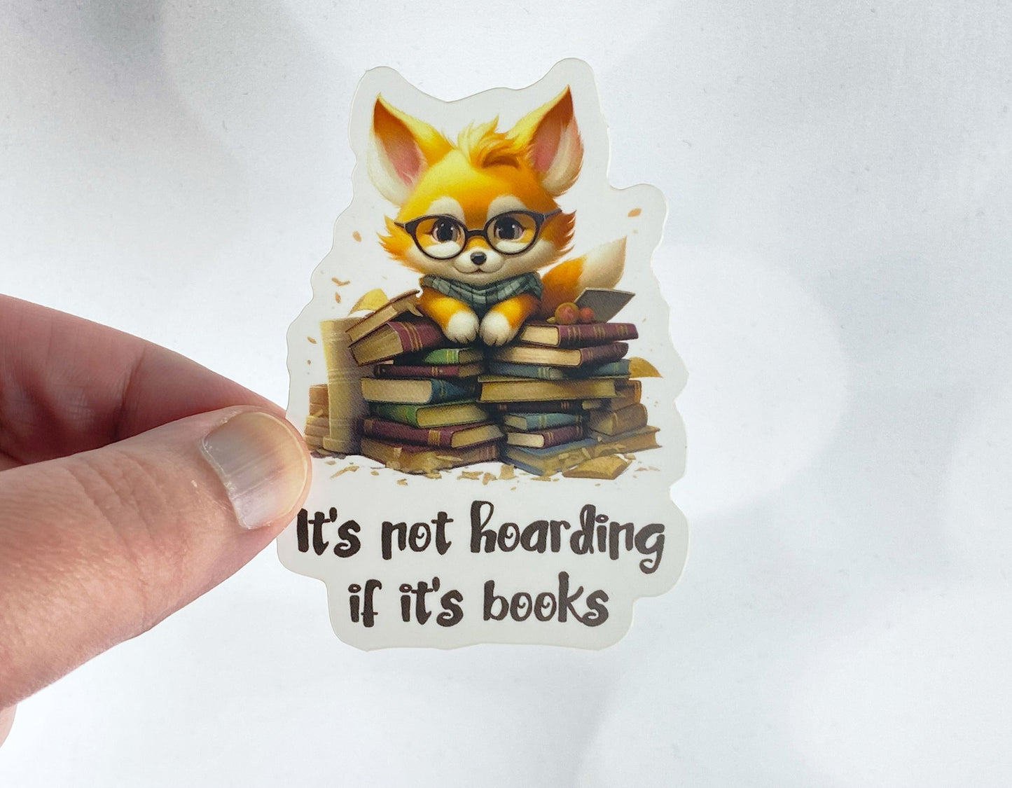 A hand holds a sticker illustration of a fox wearing glasses and a scarf, perched on a stack of books, reading "It's not hoarding if it's books," against a white background.