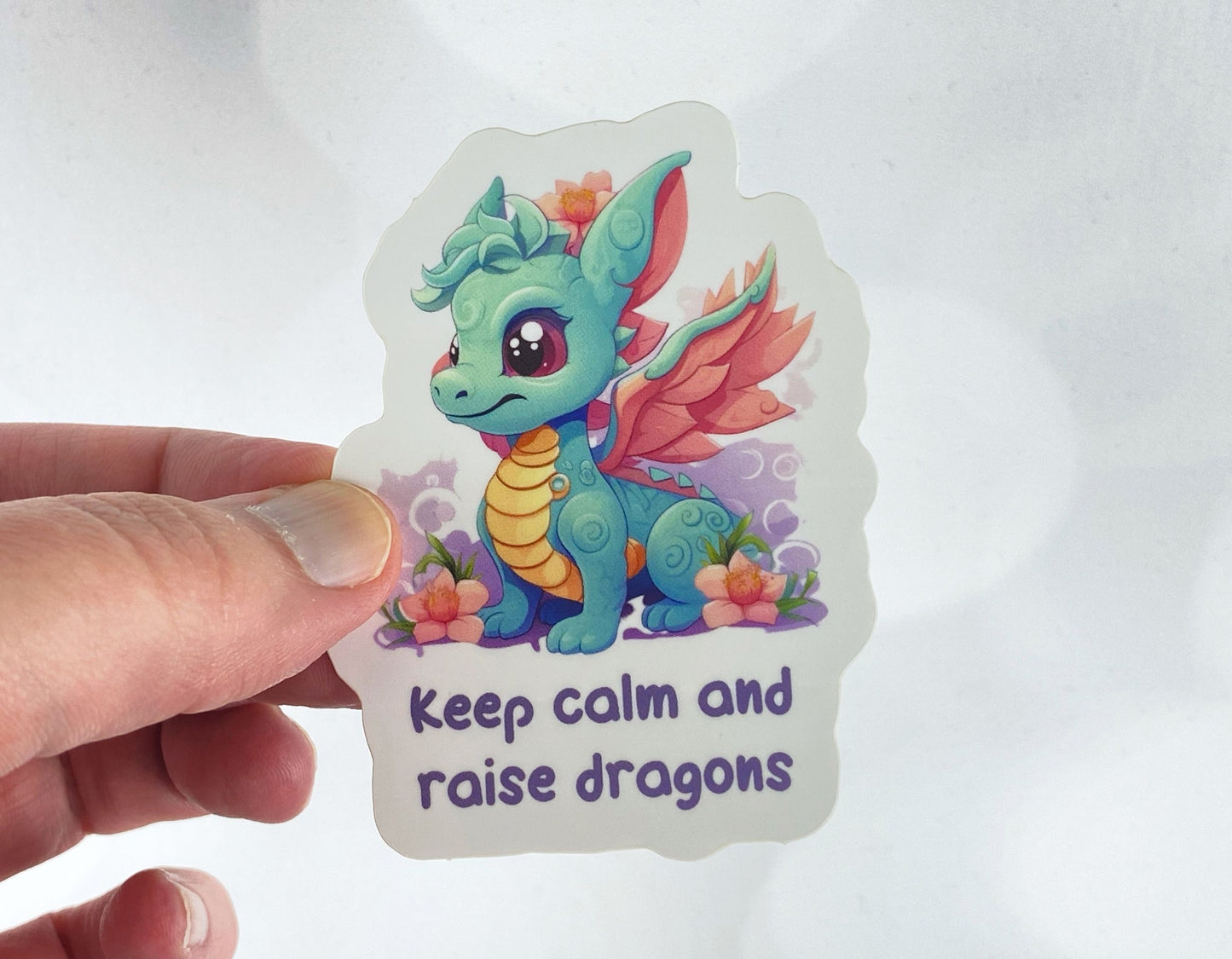 Die-cut vinyl sticker held between fingers, showing a teal baby dragon with coral wings sitting among pink flowers above the text "Keep calm and raise dragons," on a white background