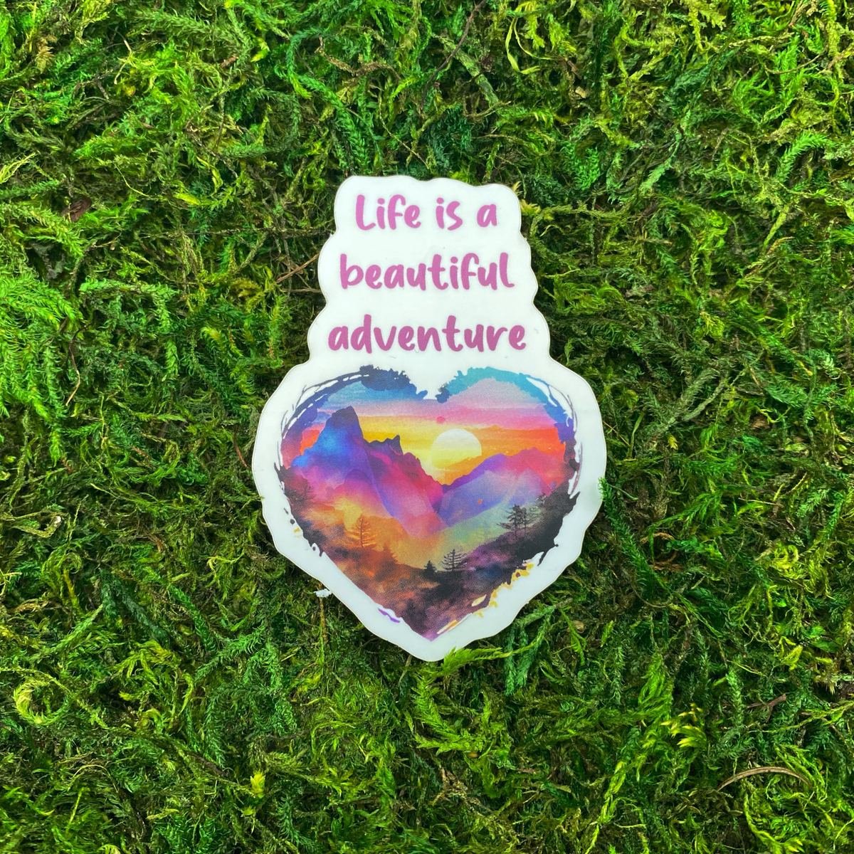 A sticker illustration of a heart-shaped watercolor mountain sunset, reading "Life is a beautiful adventure," resting on artificial moss.