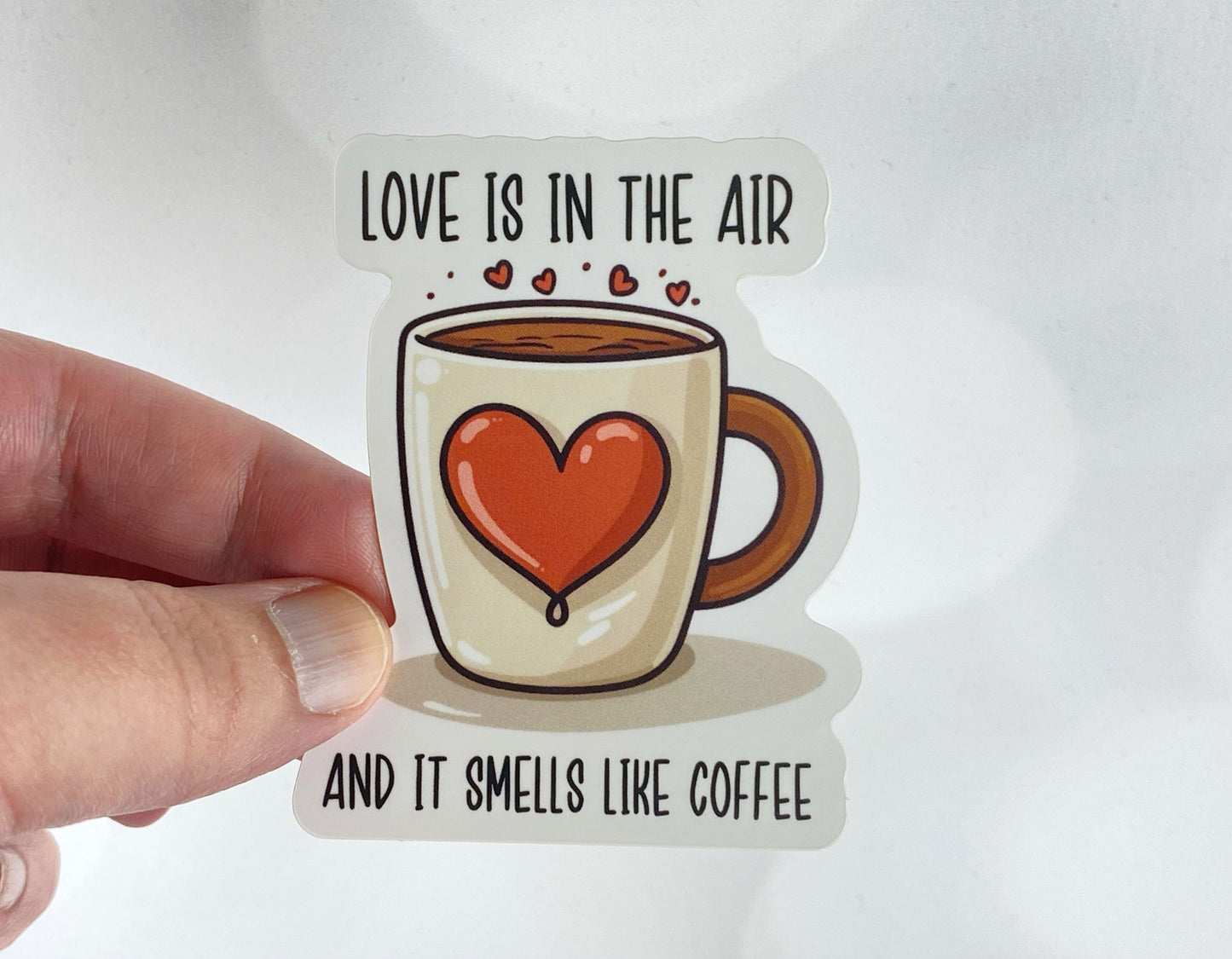 A hand holds a sticker illustration of a coffee mug with a large red heart and steam rising into small hearts, reading "Love is in the air and it smells like coffee," against a white background.