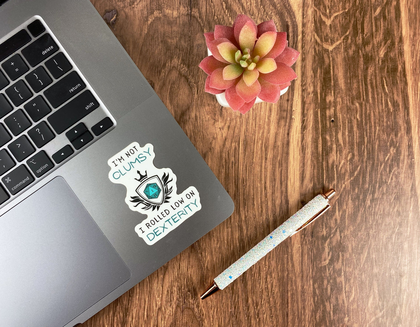 A d20 heraldic crest die-cut sticker on a laptop reading I'm not clumsy I rolled low on dexterity, next to a succulent and a glitter pen on a wood surface