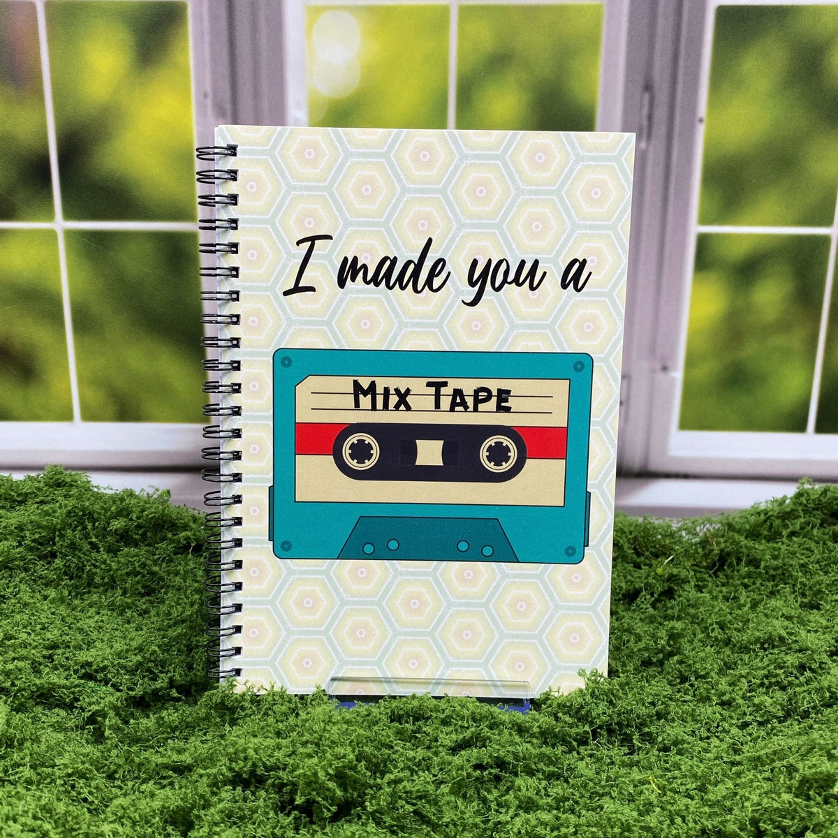 Spiral-bound notebook with a honeycomb pattern cover featuring a teal cassette tape labeled 'Mix Tape' and text reading 'I made you a,' displayed on a moss mat with a window backdrop.