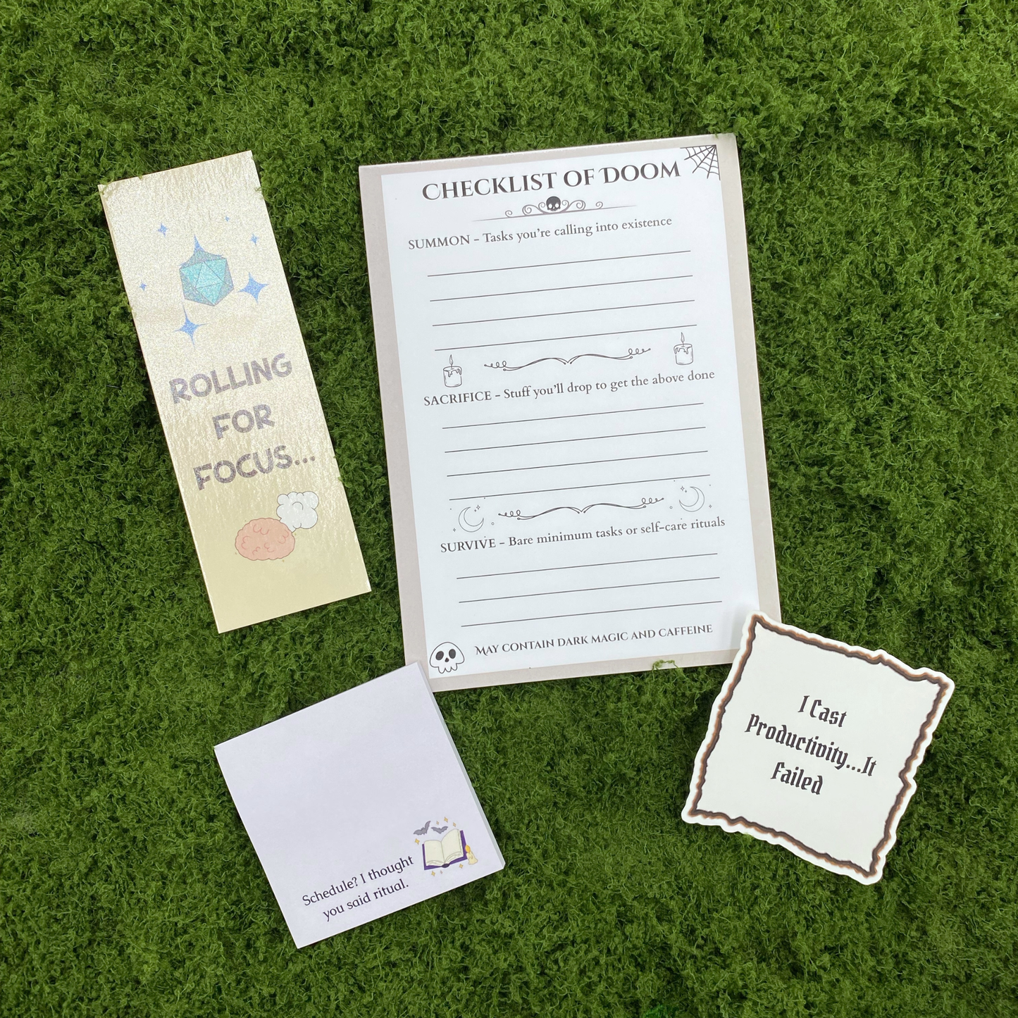 A curated gift set on artificial grass featuring a "Checklist of Doom" notepad with Summon, Sacrifice, and Survive sections, a "Rolling for Focus" bookmark with a teal d20, a sticky note reading "Schedule? I thought you said ritual," and a parchment-style sticker reading "I Cast Productivity...It Failed."