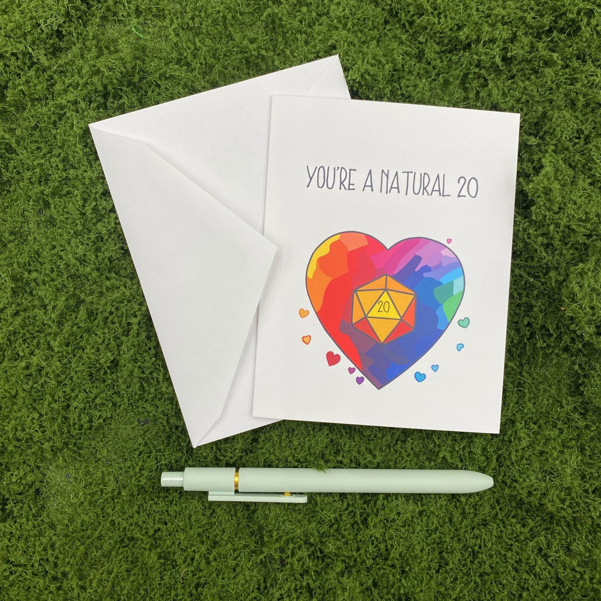 "You're a Natural 20" greeting card lying on a white envelope on grass, with a mint green pen beside it for scale.