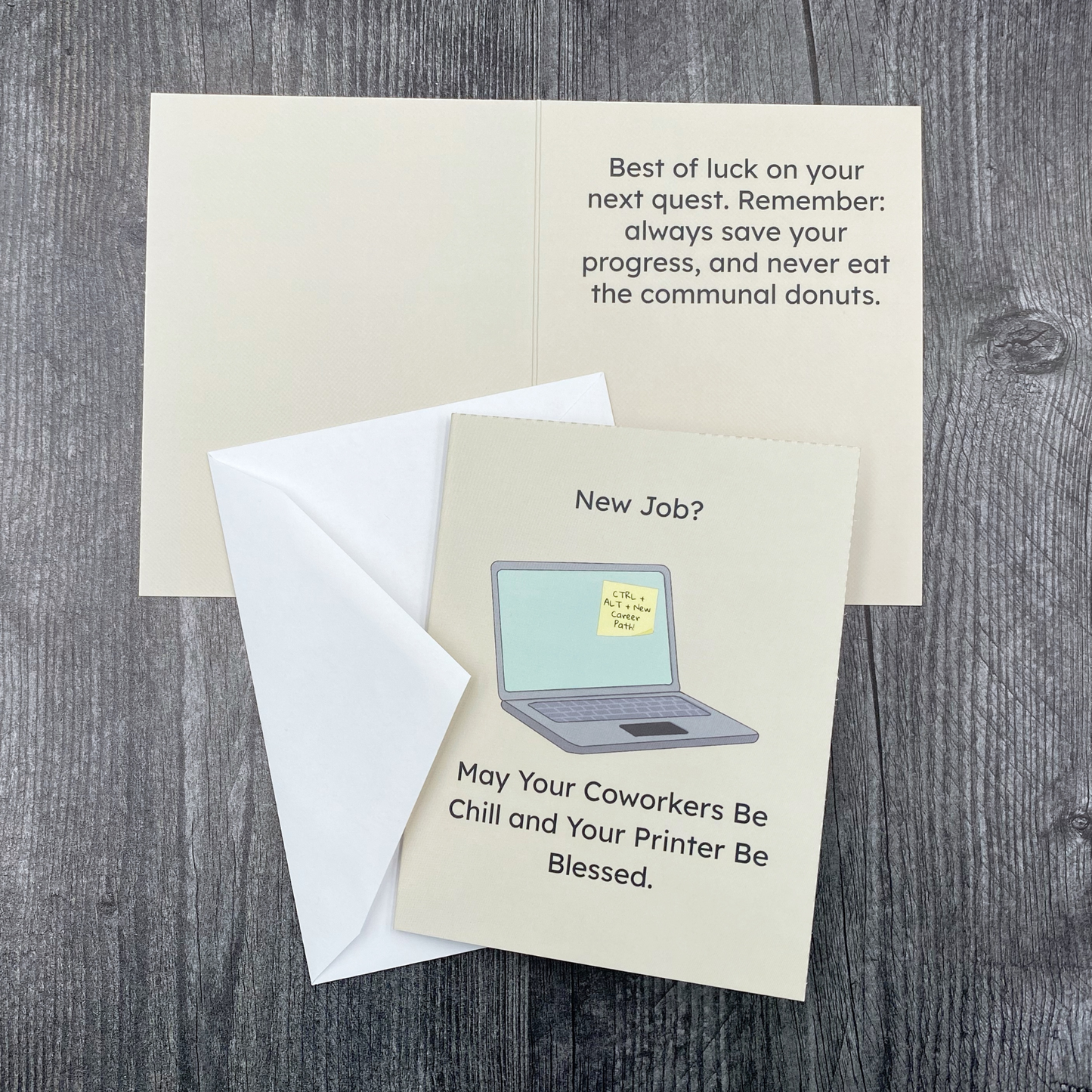 Greeting card lying open on a grey wood surface, showing the front with "New Job?" and an illustration of a laptop with a sticky note reading "CTRL + ALT + New Career Path" above "May Your Coworkers Be Chill and Your Printer Be Blessed," next to the inside message "Best of luck on your next quest. Remember: always save your progress, and never eat the communal donuts," with the envelope tucked beneath the front cover.