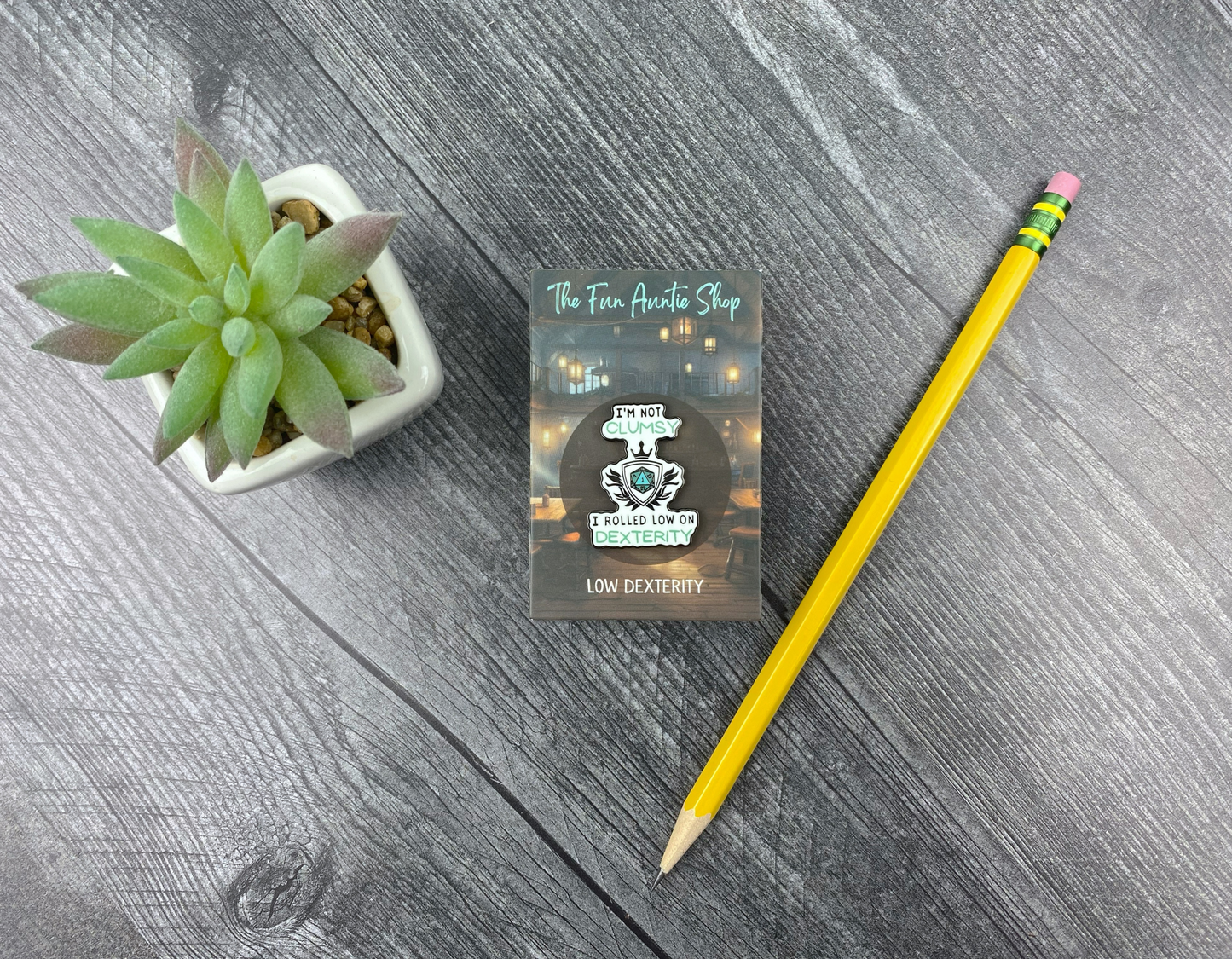Enamel pin shaped like a shield crest with a d20 in the center, reading "I'm not clumsy, I rolled low on dexterity," on a backing card next to a succulent and pencil on a gray wood background