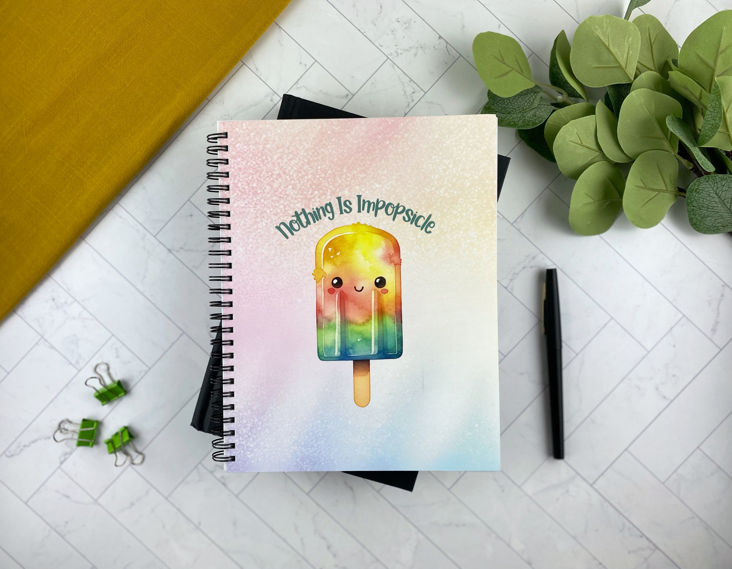 Spiral notebook with a pastel cover featuring a smiling rainbow popsicle and the text "Nothing is impopsicle," resting on a marble surface next to a plant, pen, and binder clips