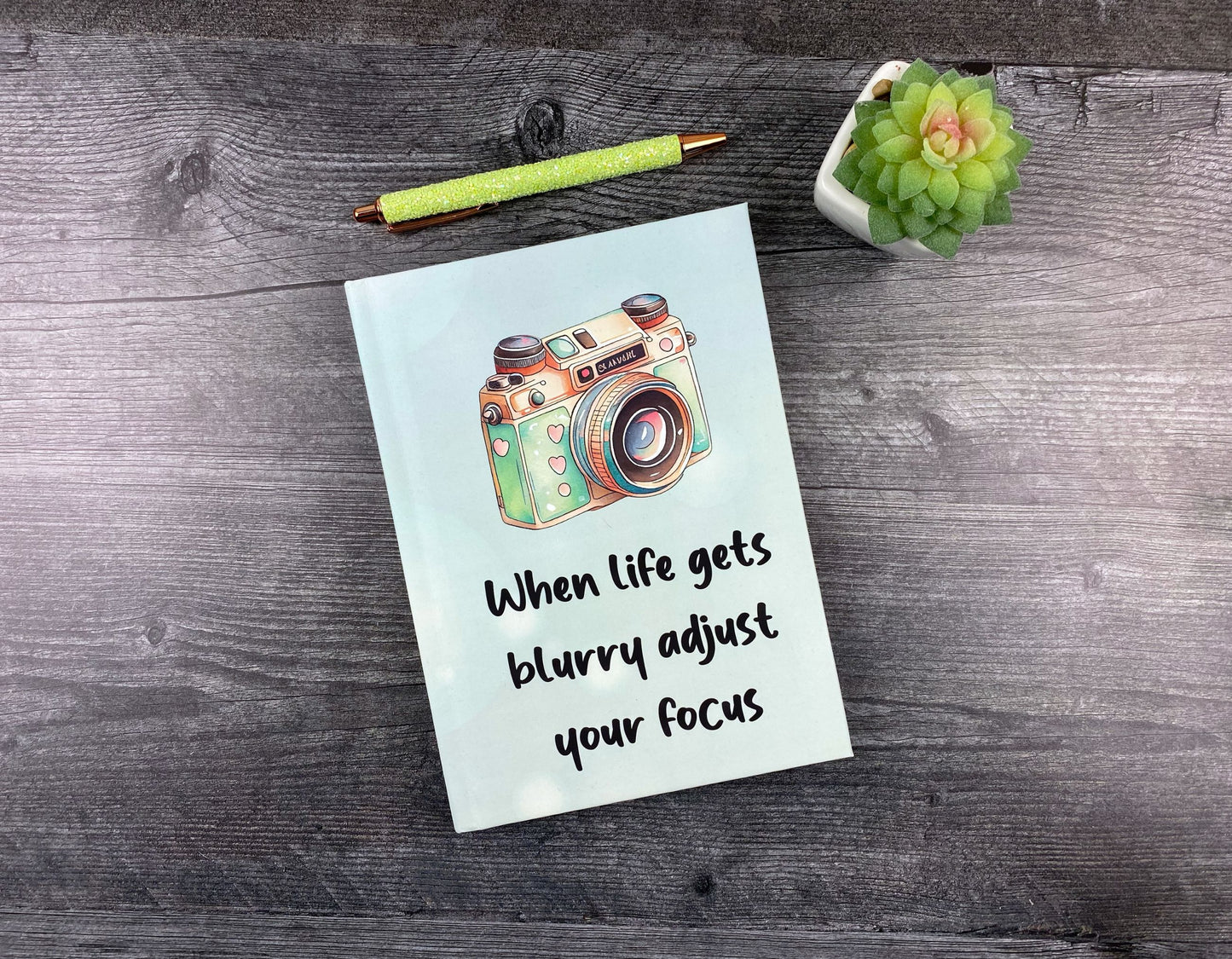 Hardcover journal in pale blue with an illustrated vintage camera and the words "When life gets blurry, adjust your focus," resting on a grey wood surface next to a glitter pen and small succulent.