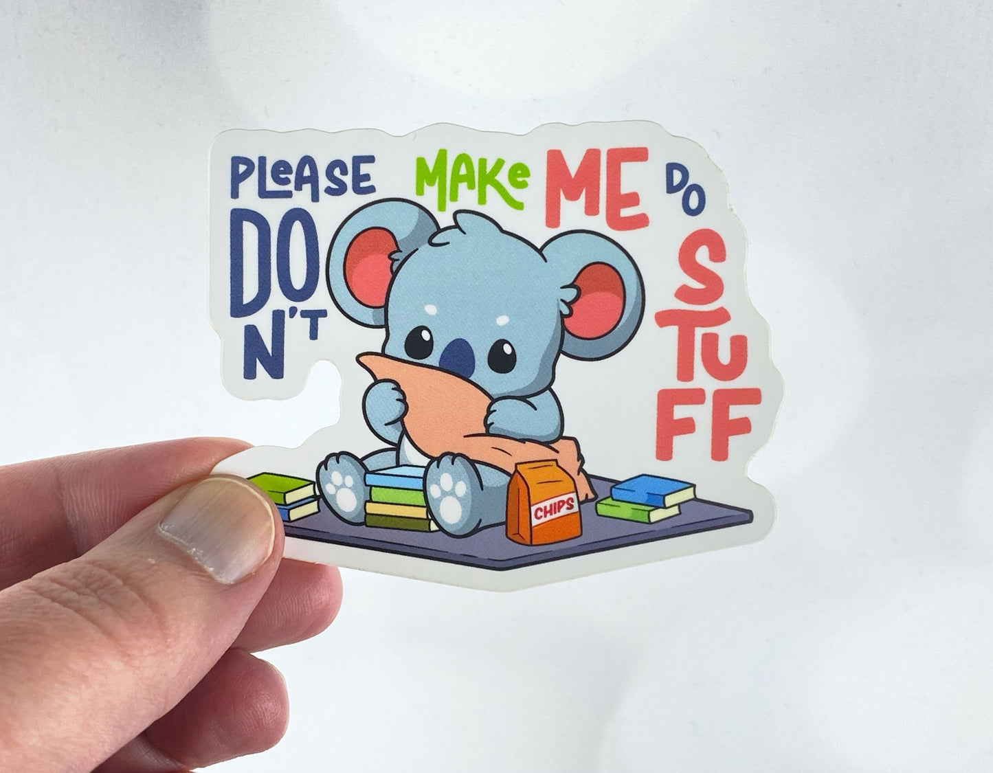 Die-cut vinyl sticker held between fingers, showing a koala wrapped in a blanket surrounded by books and a bag of chips, with the text "Please don't make me do stuff," on a white background