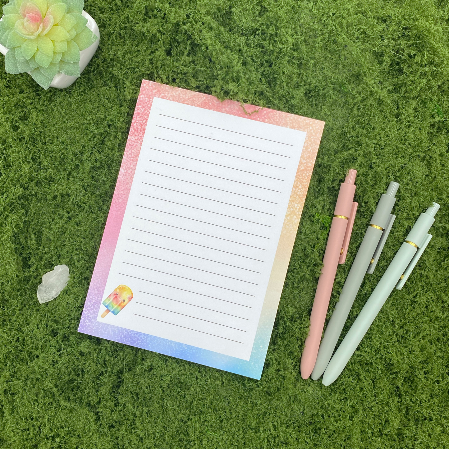 Lined notepad with a pastel rainbow watercolor border and a smiling popsicle illustration in the corner, resting on artificial grass next to a crystal, small succulent, and three pastel gel pens.