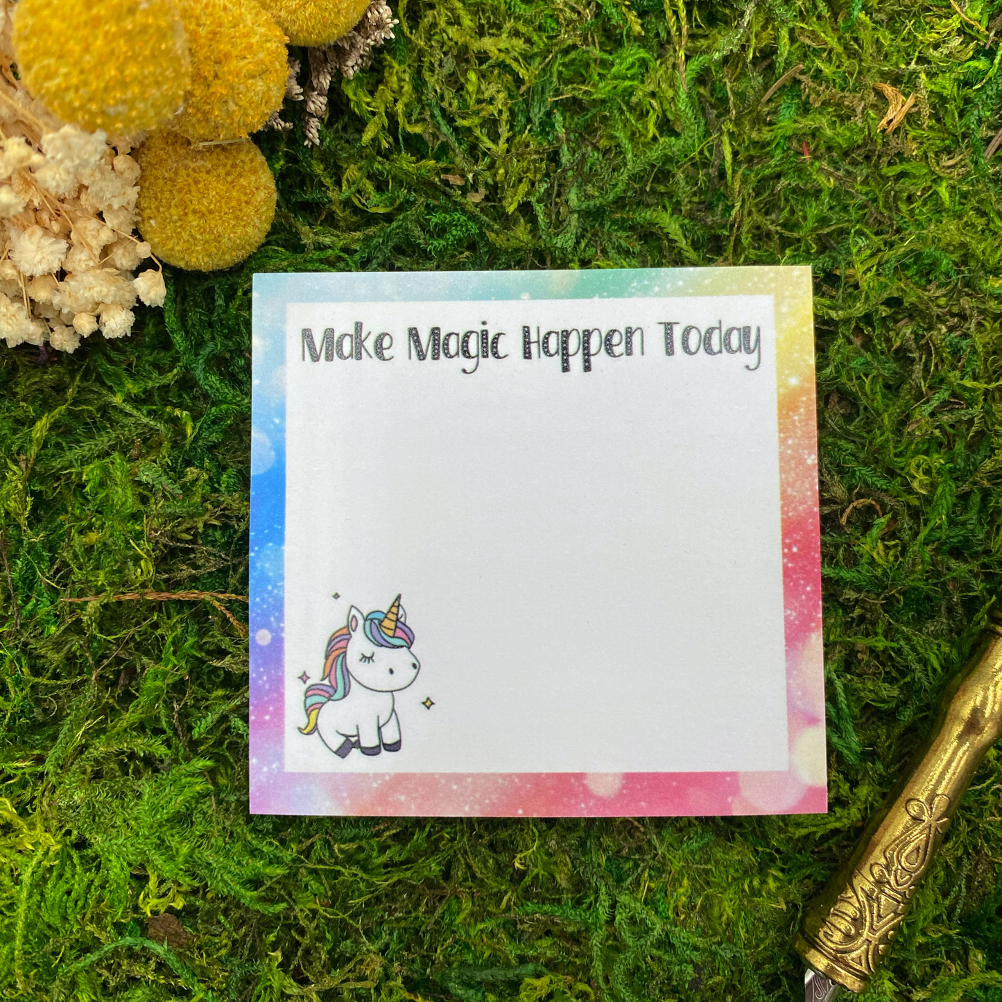 Square sticky note pad with a rainbow galaxy swirl border, a small unicorn illustration, and the words "Make Magic Happen Today," resting on moss next to dried yellow and white flowers and a gold quill pen.