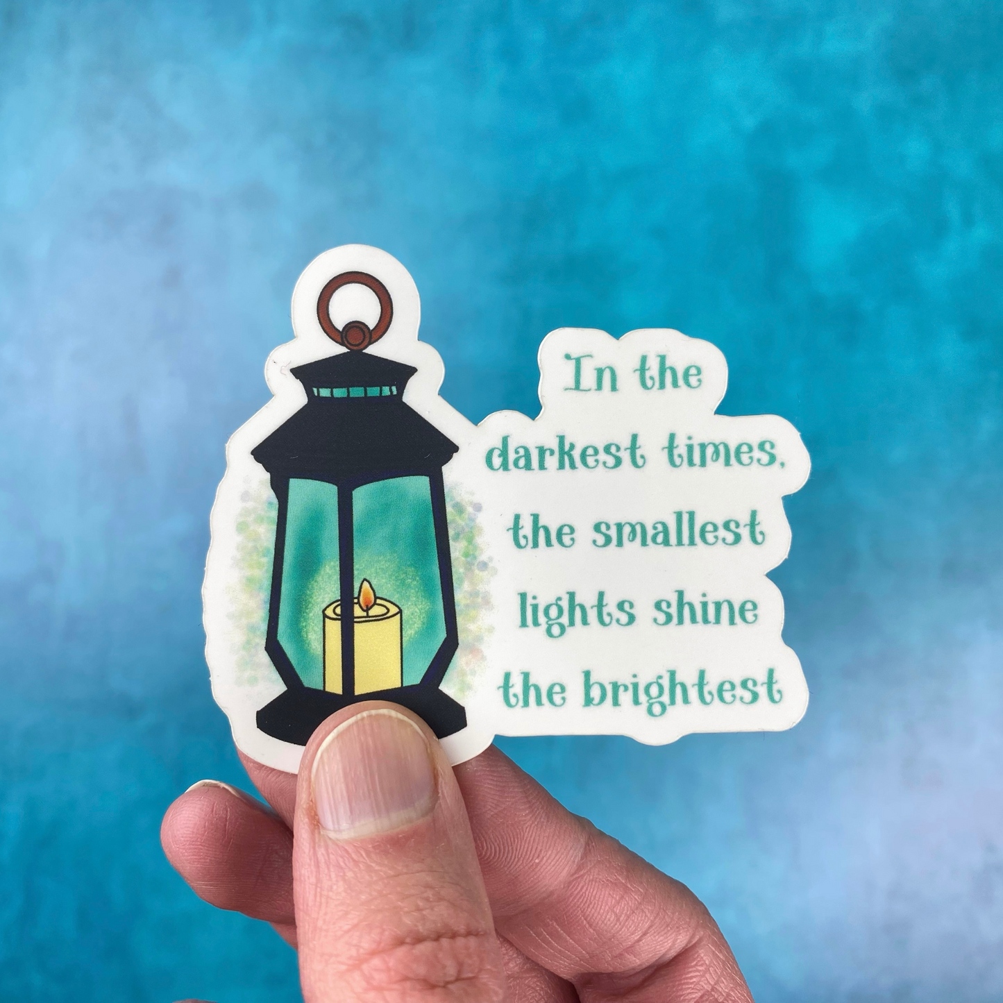 Die-cut vinyl sticker showing a black lantern with a glowing candle inside, with the text "In the darkest times, the smallest lights shine the brightest," on a teal background