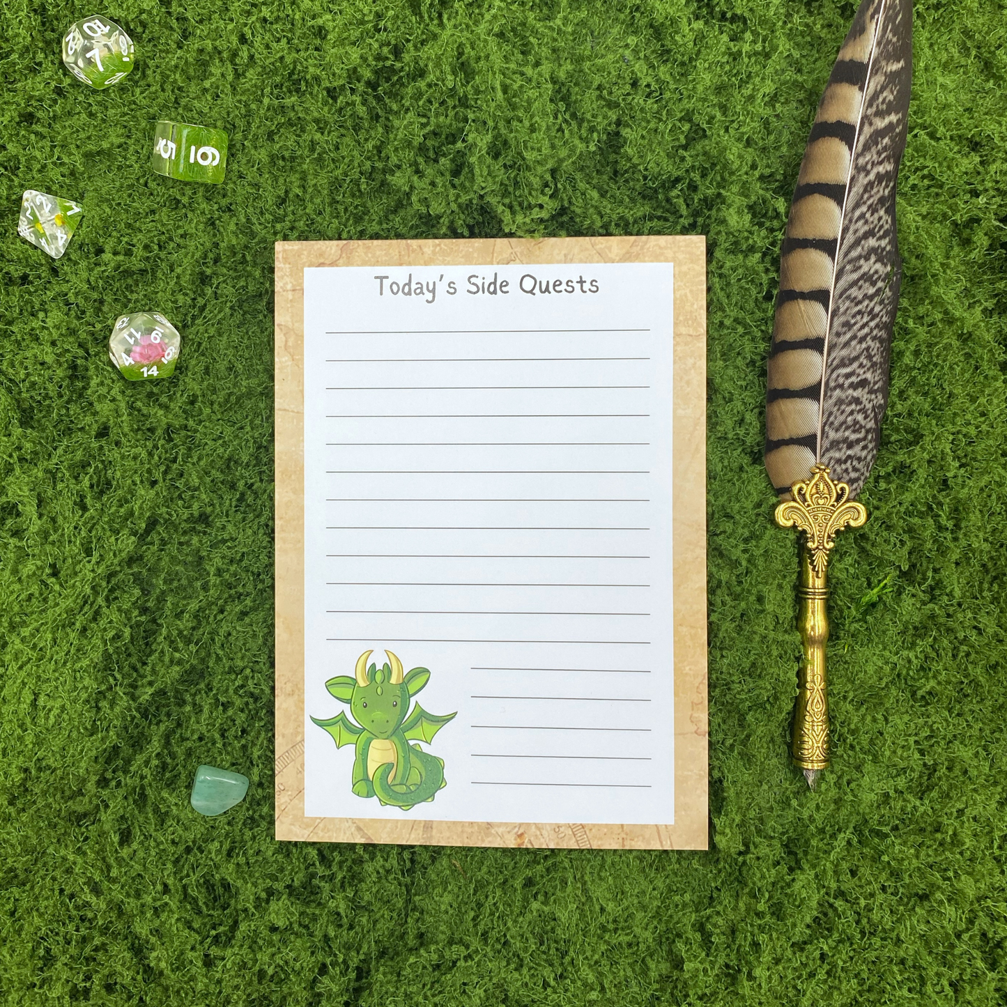 Lined notepad on a parchment-textured border headed "Today's Side Quests," with a small green dragon illustration in the corner, resting on artificial grass next to dice, a crystal, and a gold quill pen.
