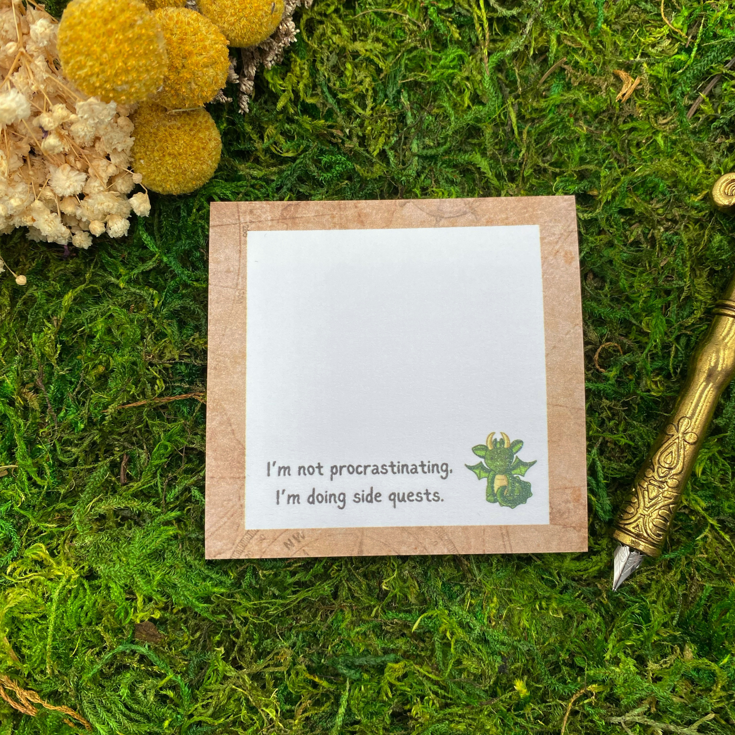 Square sticky note pad with a tan map-textured border, a small green dragon illustration, and the words "I'm not procrastinating, I'm doing side quests," resting on moss next to dried yellow and white flowers and a gold quill pen.