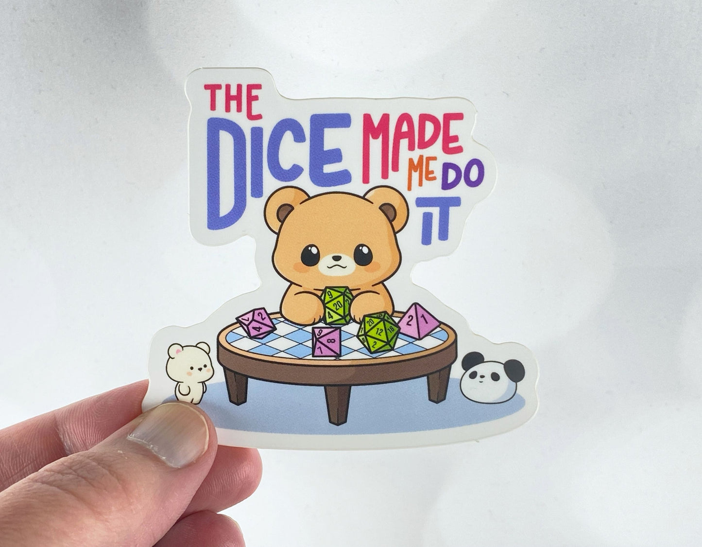 Die-cut vinyl sticker held between fingers, showing a bear rolling dice at a round table with a bear and panda nearby, above the text "The dice made me do it," on a white background
