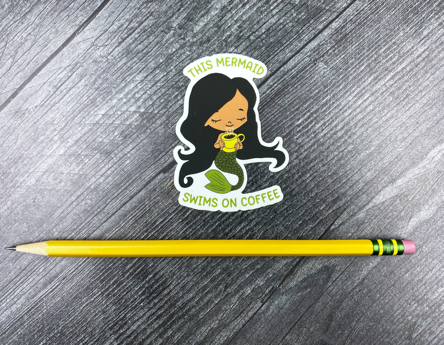 Die-cut vinyl sticker showing a dark-haired mermaid with a green tail holding a steaming yellow mug, with the text "This mermaid swims on coffee," on a gray wood background next to a yellow pencil