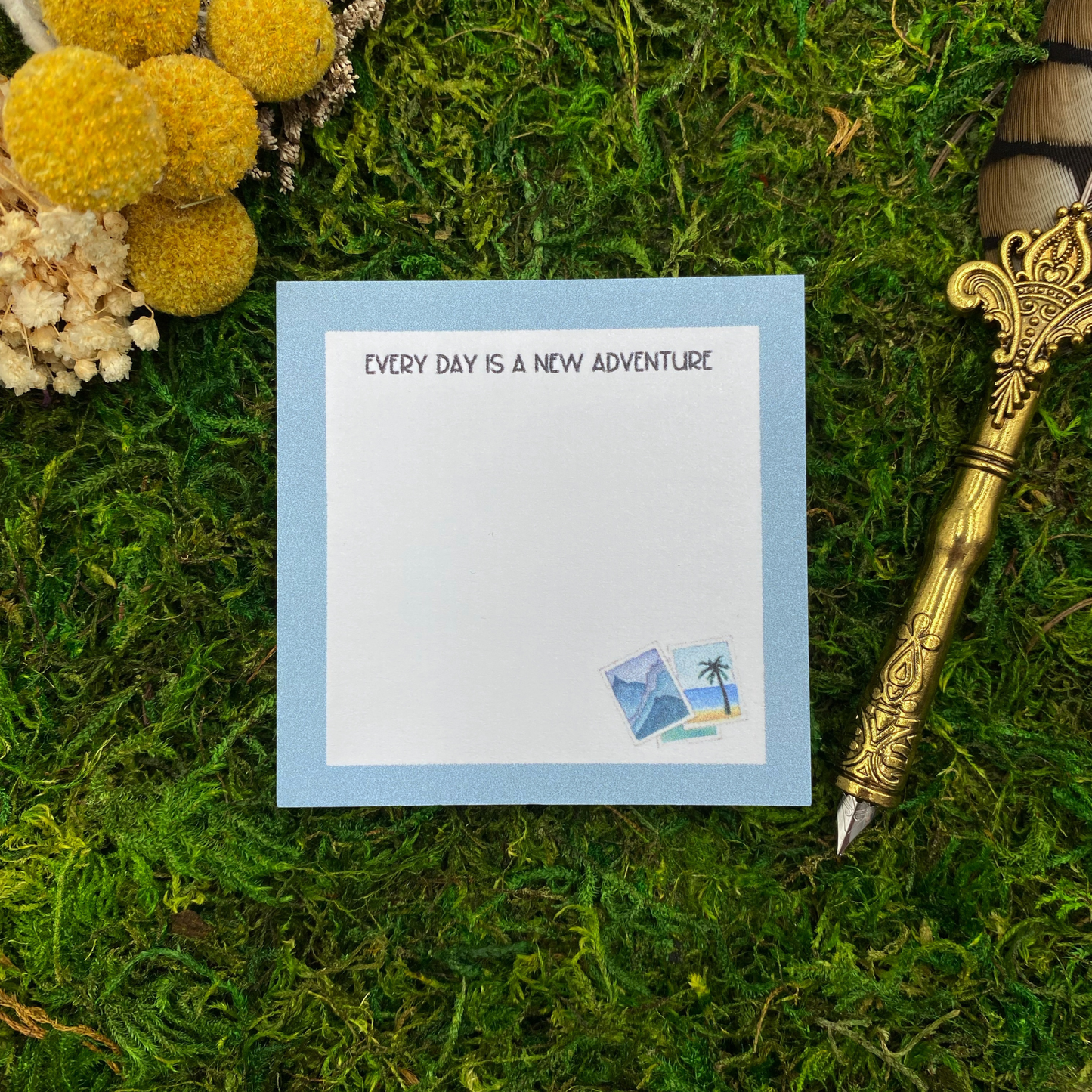 Square sticky note pad with a light blue border, small illustrated travel photos of mountains and a palm tree beach, and the words "Every day is a new adventure," resting on moss next to dried yellow and white flowers and a gold quill pen.