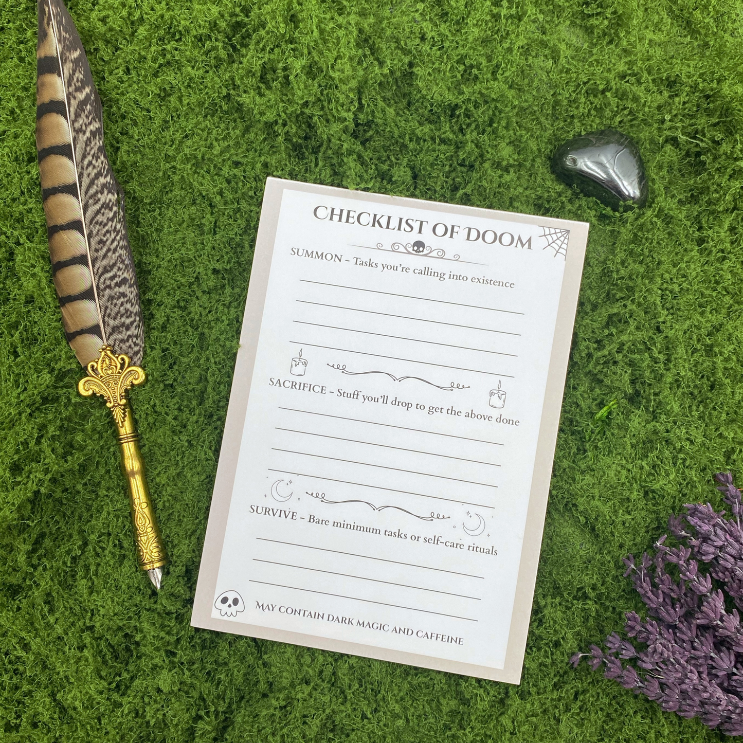Notepad headed "Checklist of Doom" with three sections labeled Summon, Sacrifice, and Survive, decorated with a spiderweb, skull, candles, and crescent moons, resting on artificial grass next to a gold quill pen, an obsidian stone, and dried lavender.